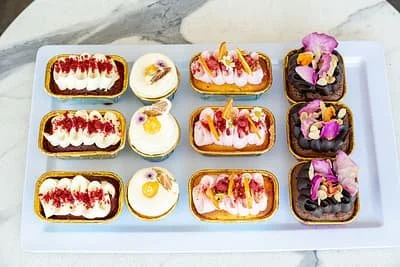 A white rectangular platter holding twelve small decorated desserts with various toppings and colors.