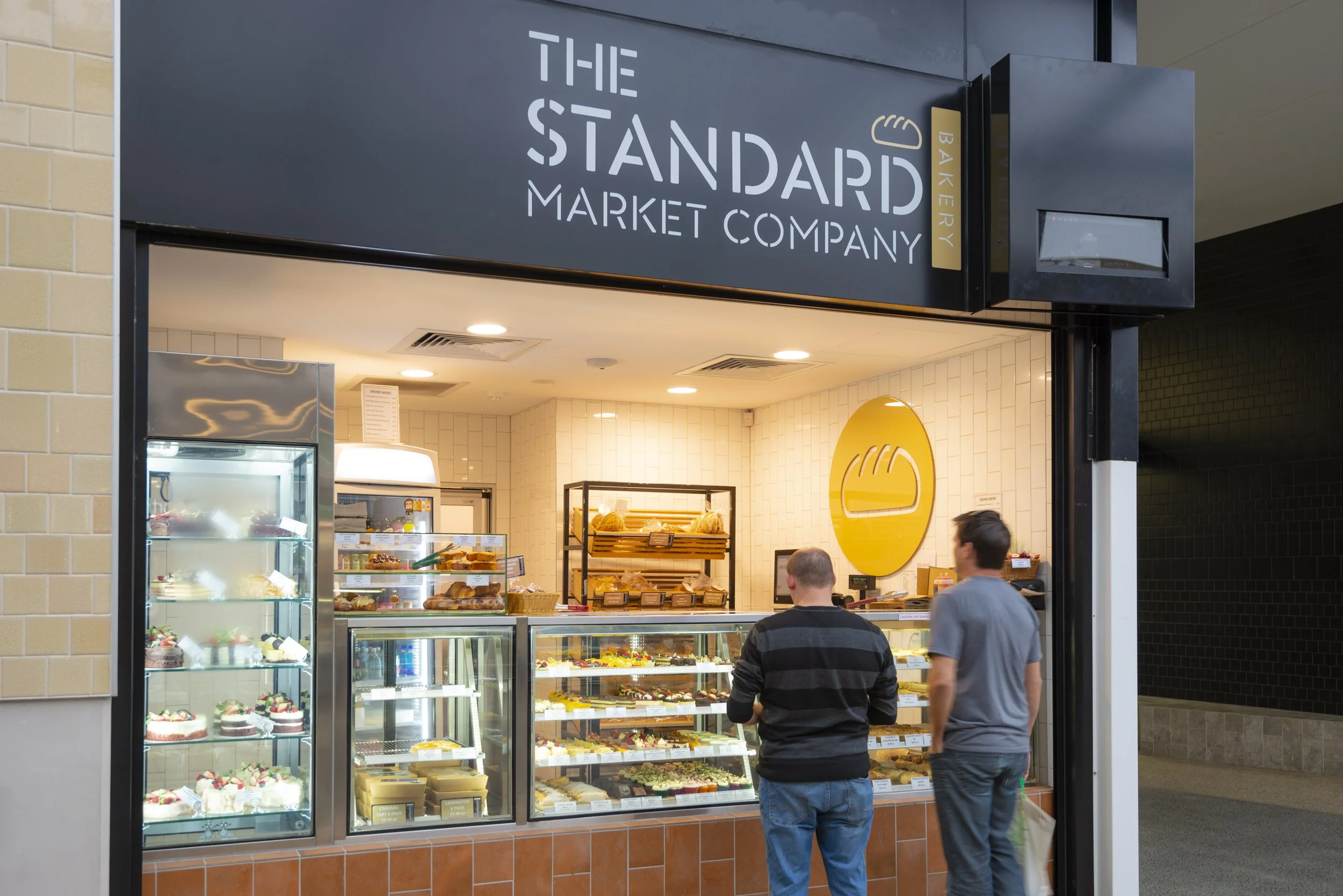 View of a bakery inside a market called 'The Standard Market Company', with display cases filled with cakes, pastries, and baked goods, and two people standing in front of the display selecting items.