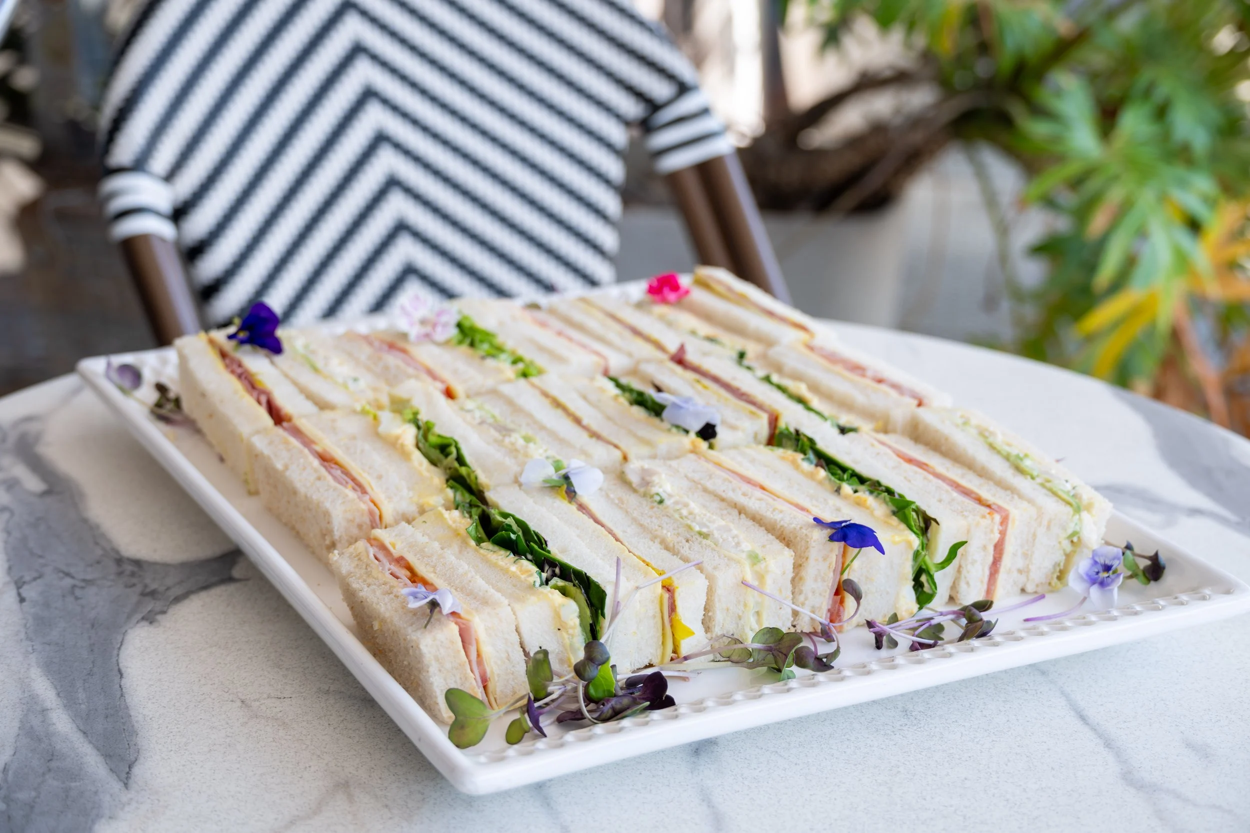 Plate of assorted sandwiches with lettuce, tomato, and various fillings, decorated with edible flowers on a marble table.