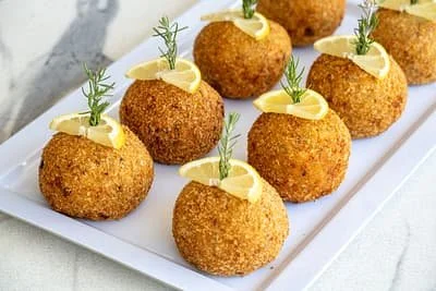 Fried arancini balls garnished with lemon slices and rosemary on a white platter.