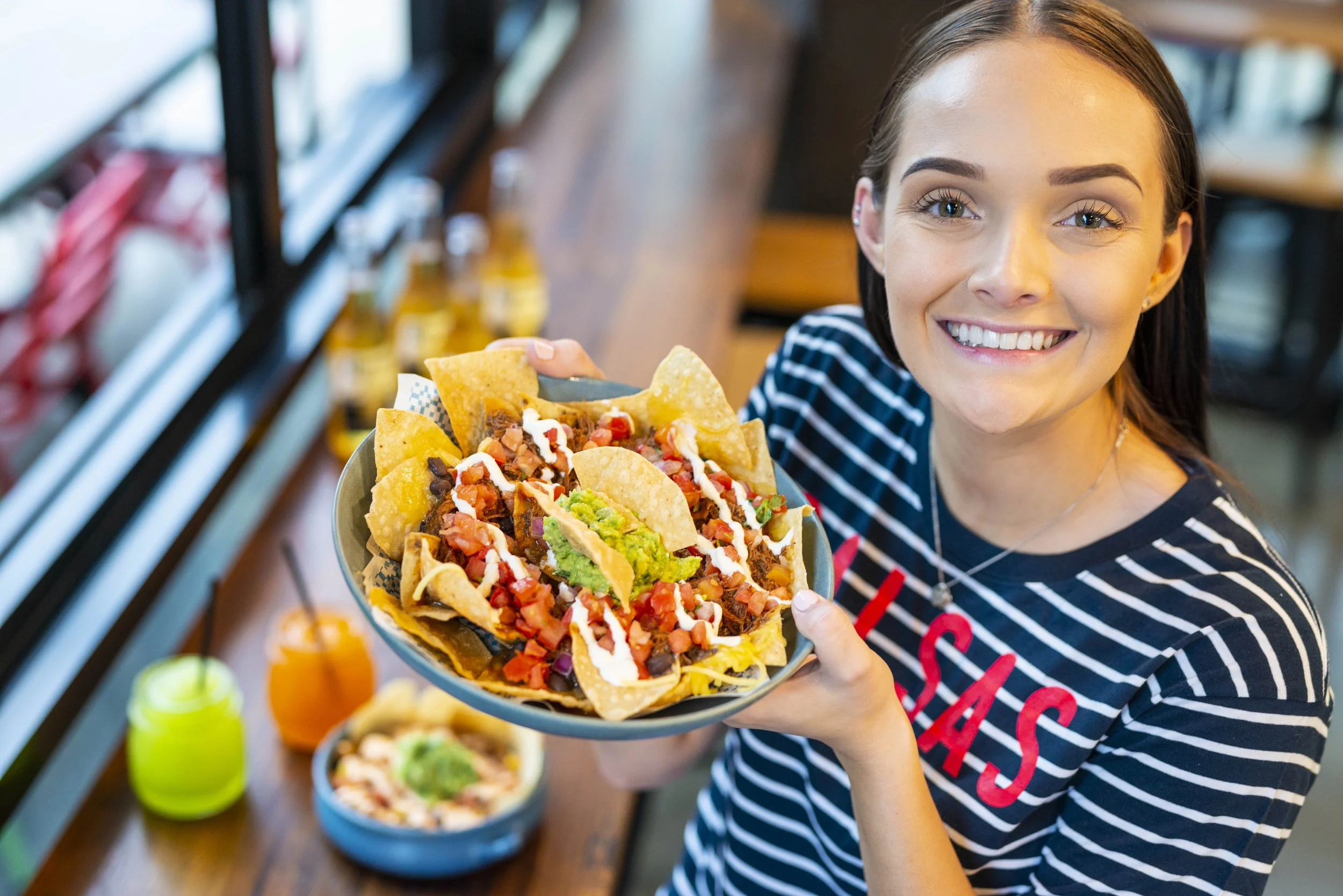 A young woman with dark hair and blue eyes smiling and holding a plate of nachos topped with cheese, tomatoes, sour cream, guacamole, and other toppings inside a restaurant.