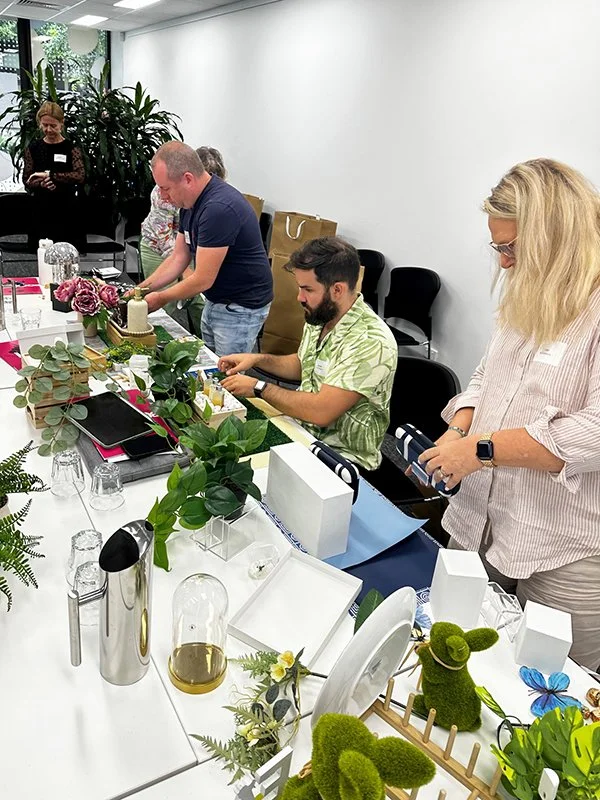 People gathered around a table with plants, flowers, and gift boxes, participating in a workshop.