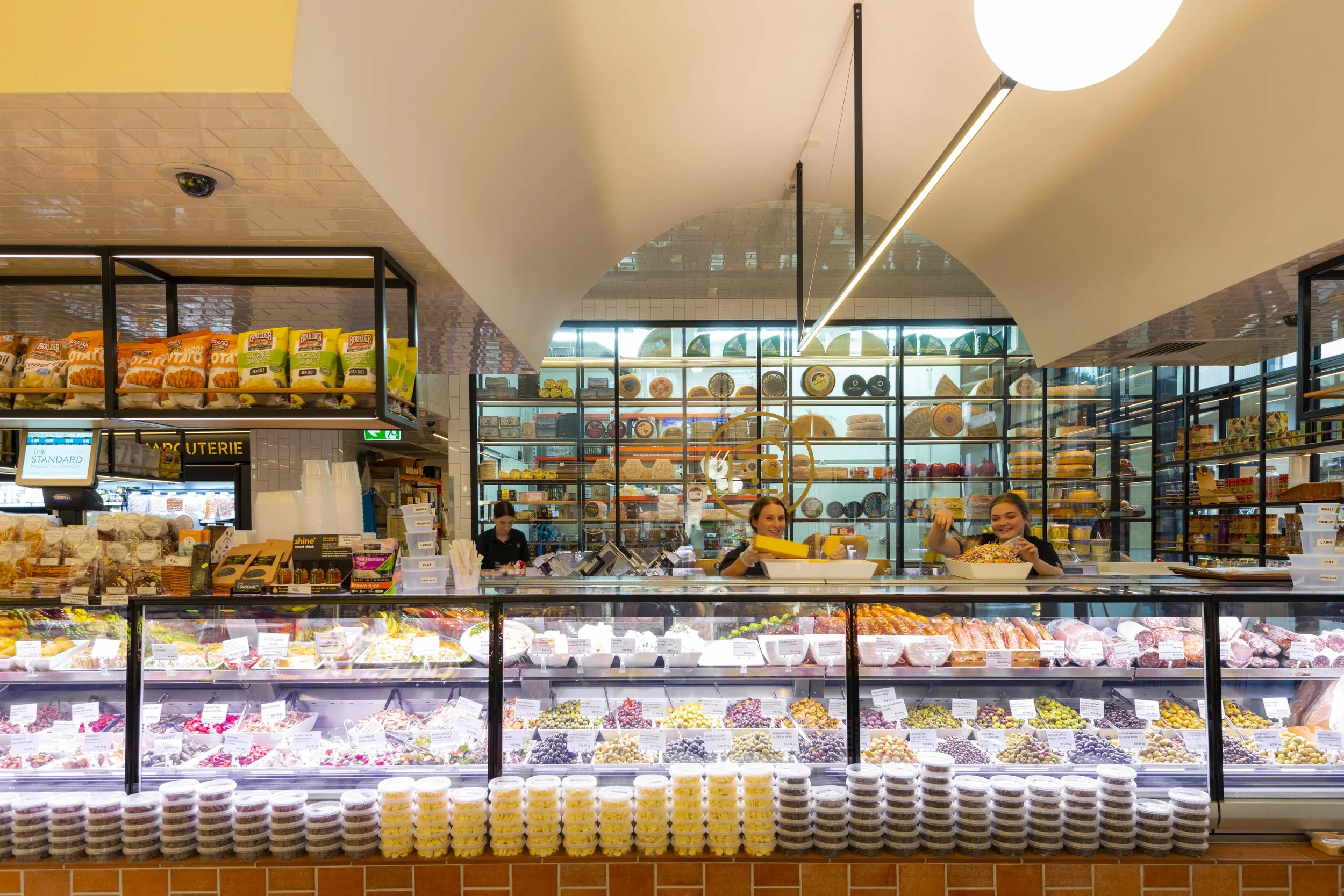 Cheese and meat deli counter with employees behind the counter in a grocery store, shelves with cheese wheels in the background, and various packaged products on top.