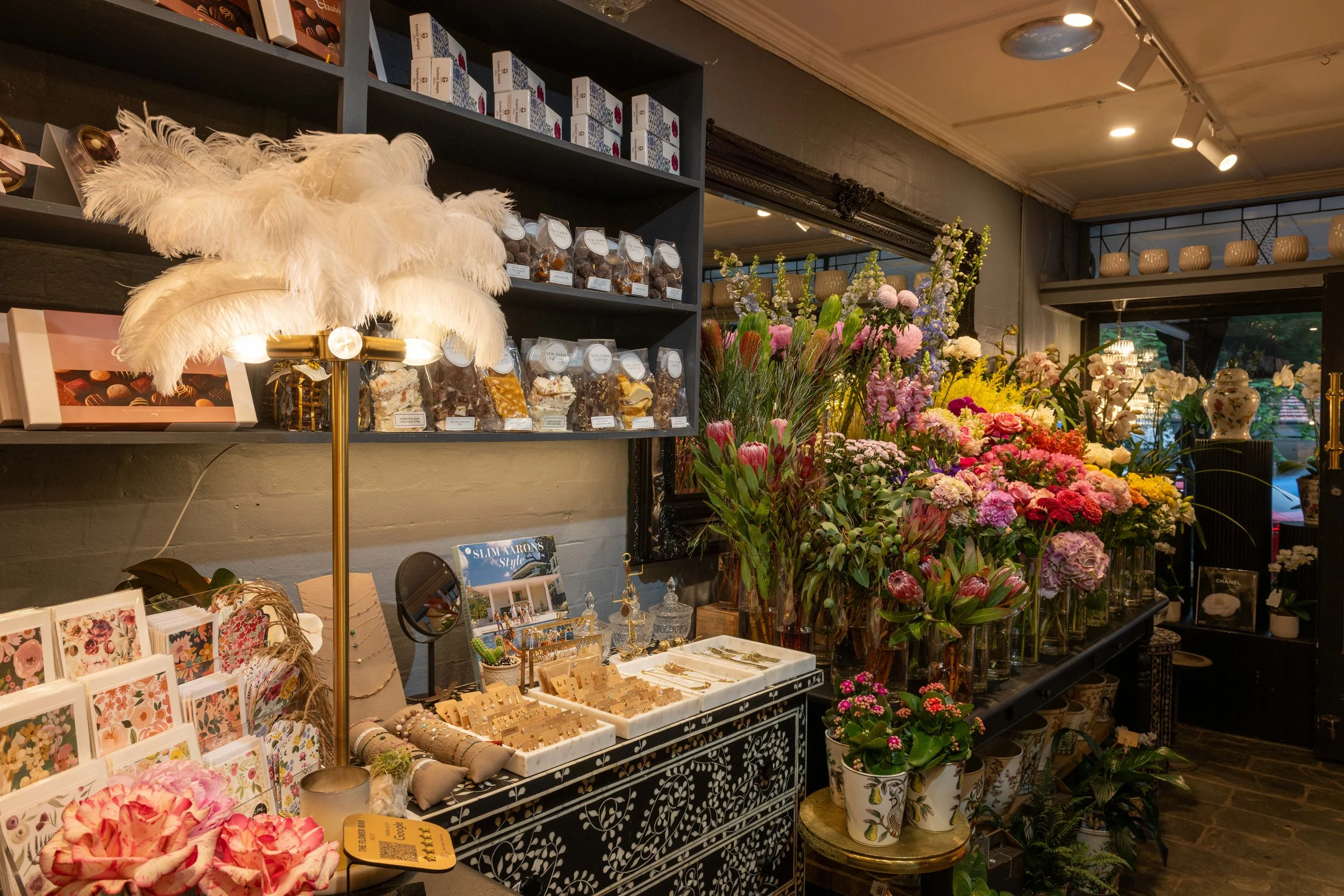 Inside a flower shop with various colorful flowers and plants on display, jewelry and decorative items on tables, and a lamp with white feathers.