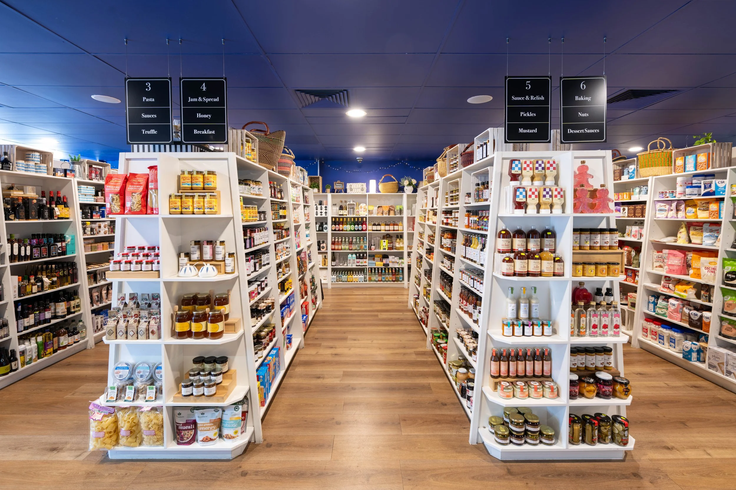 View of grocery store shelves with various products, signs overhead indicating different sections such as pasta, sauces, jam, spreads, and baked goods.