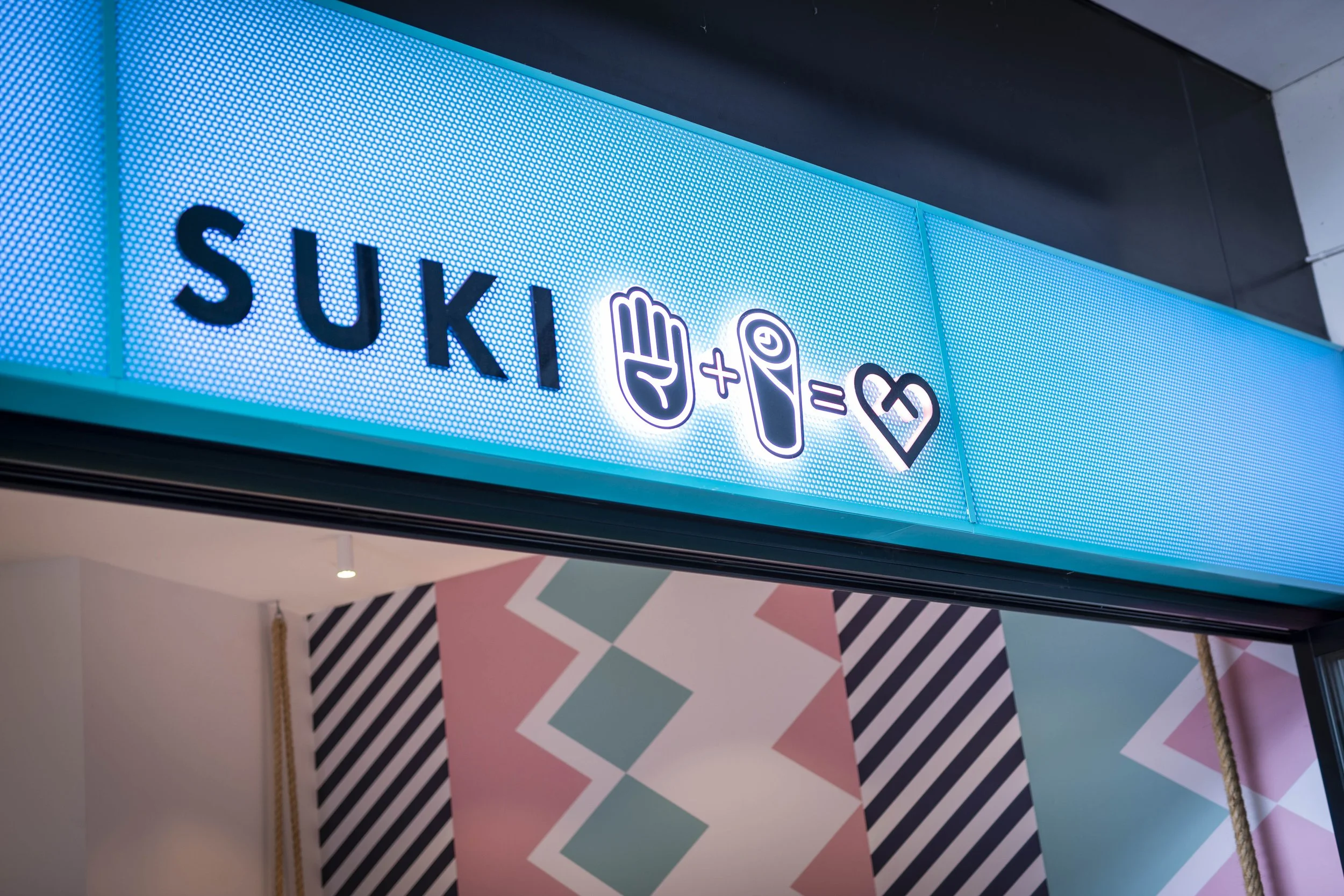 A blue illuminated sign with the name 'SUKI' followed by a playful equation using symbols: a hand with a raised thumb plus a rolled sushi roll equals a heart shape.