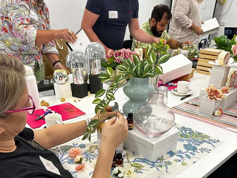 People gathered around a decorated table with flowers, vases, and craft supplies, participating in a workshop.