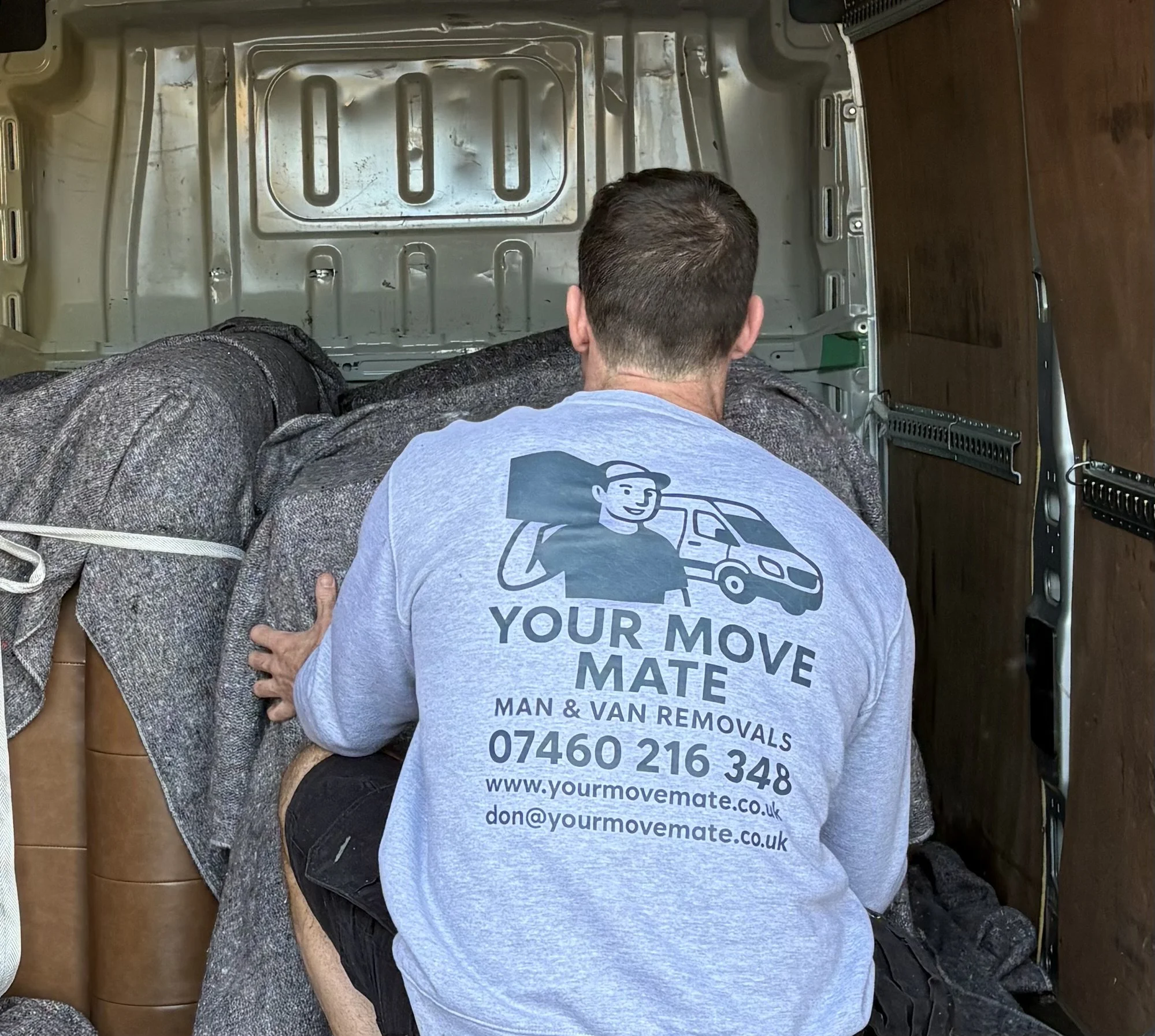 Don wearing branded your move mate jumper, kneeling and lifting a large piece of furniture inside a moving van.