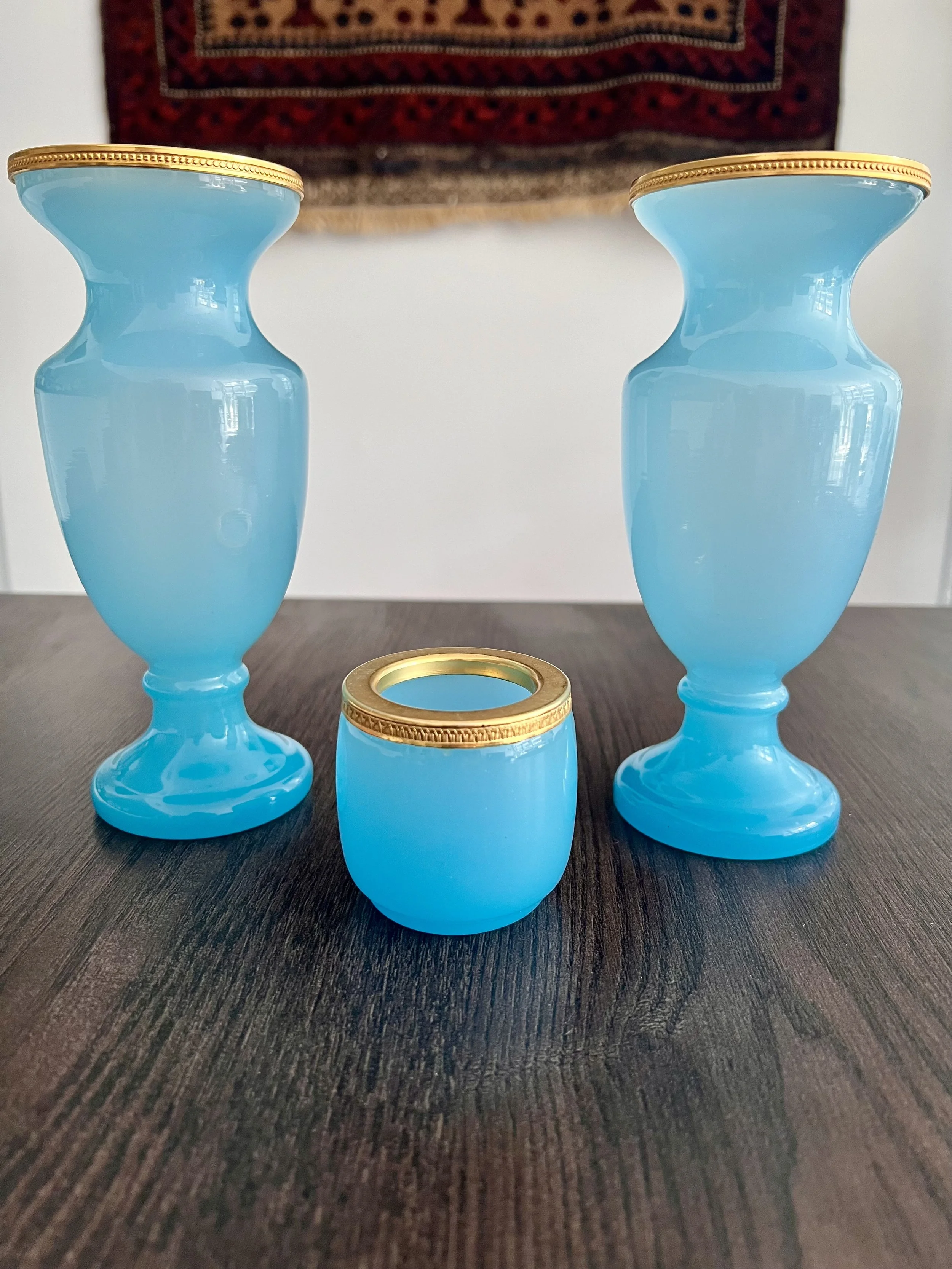A three-piece antique garniture set made of turquoise blue opaline glass with ornate gilded gold rims, featuring two matching baluster vases and a central cup on a dark wood table.