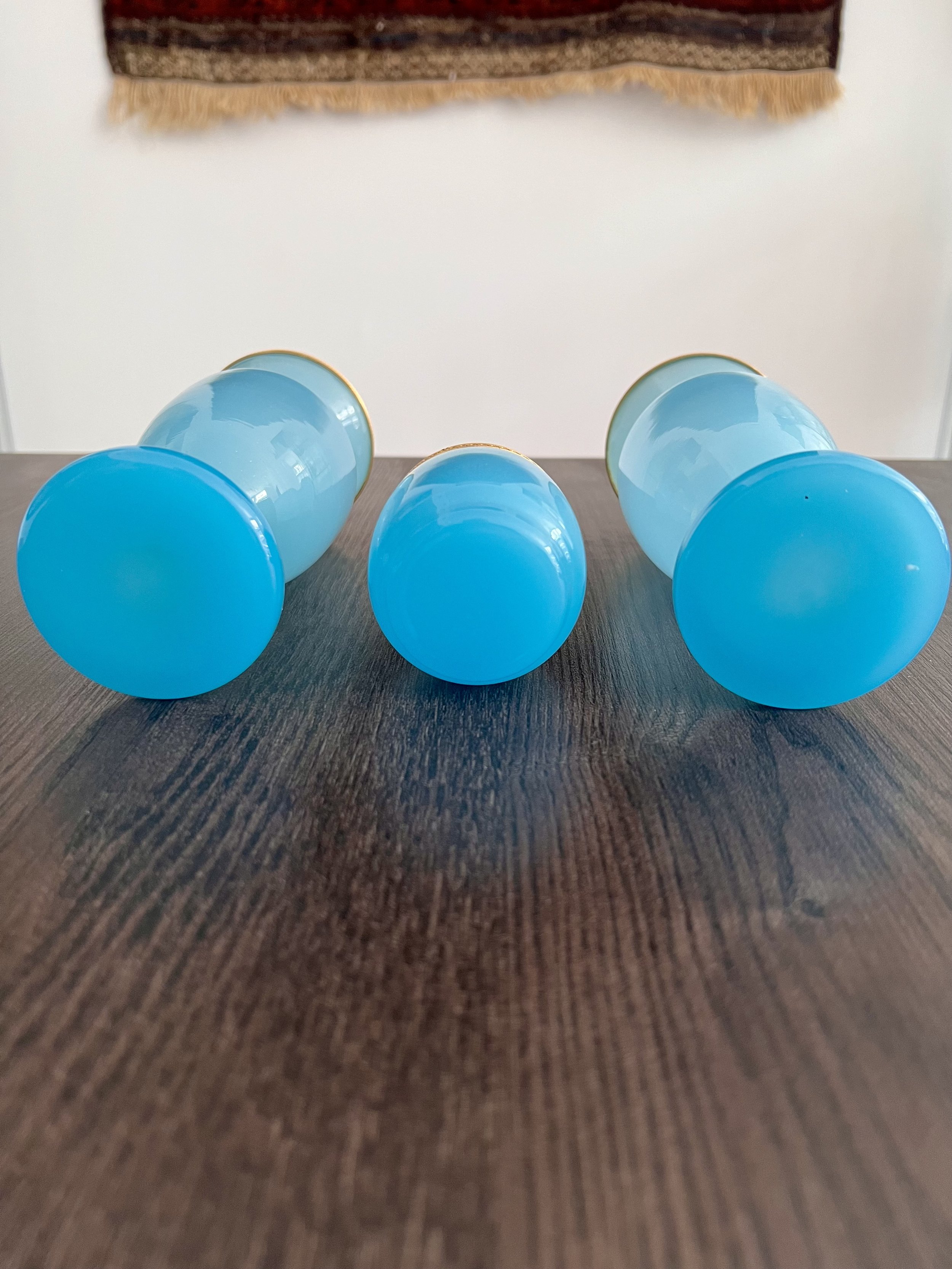 A three-piece antique garniture set made of turquoise blue opaline glass with ornate gilded gold rims, featuring two matching baluster vases and a central cup on a dark wood table.