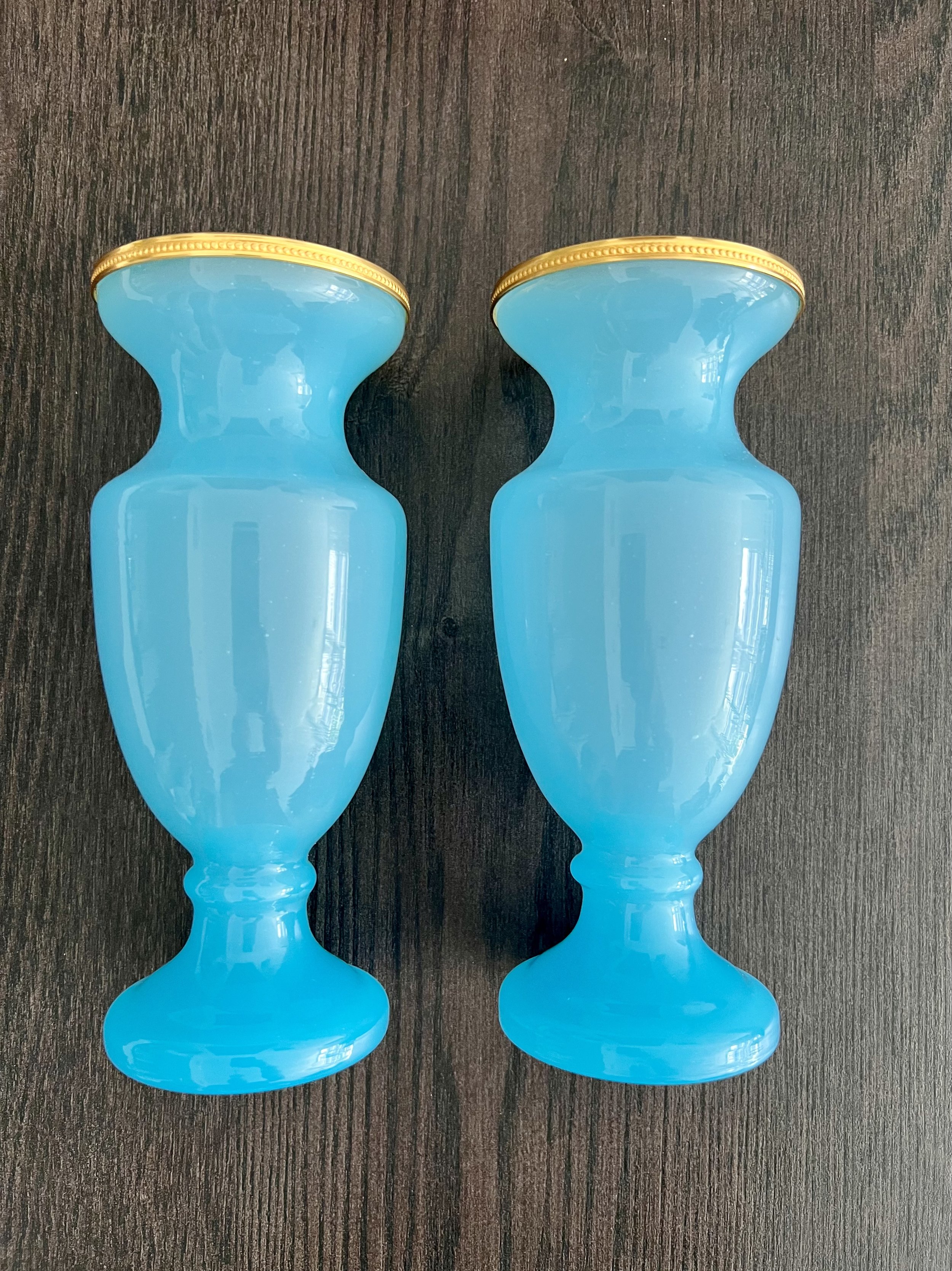 A three-piece antique garniture set made of turquoise blue opaline glass with ornate gilded gold rims, featuring two matching baluster vases and a central cup on a dark wood table.