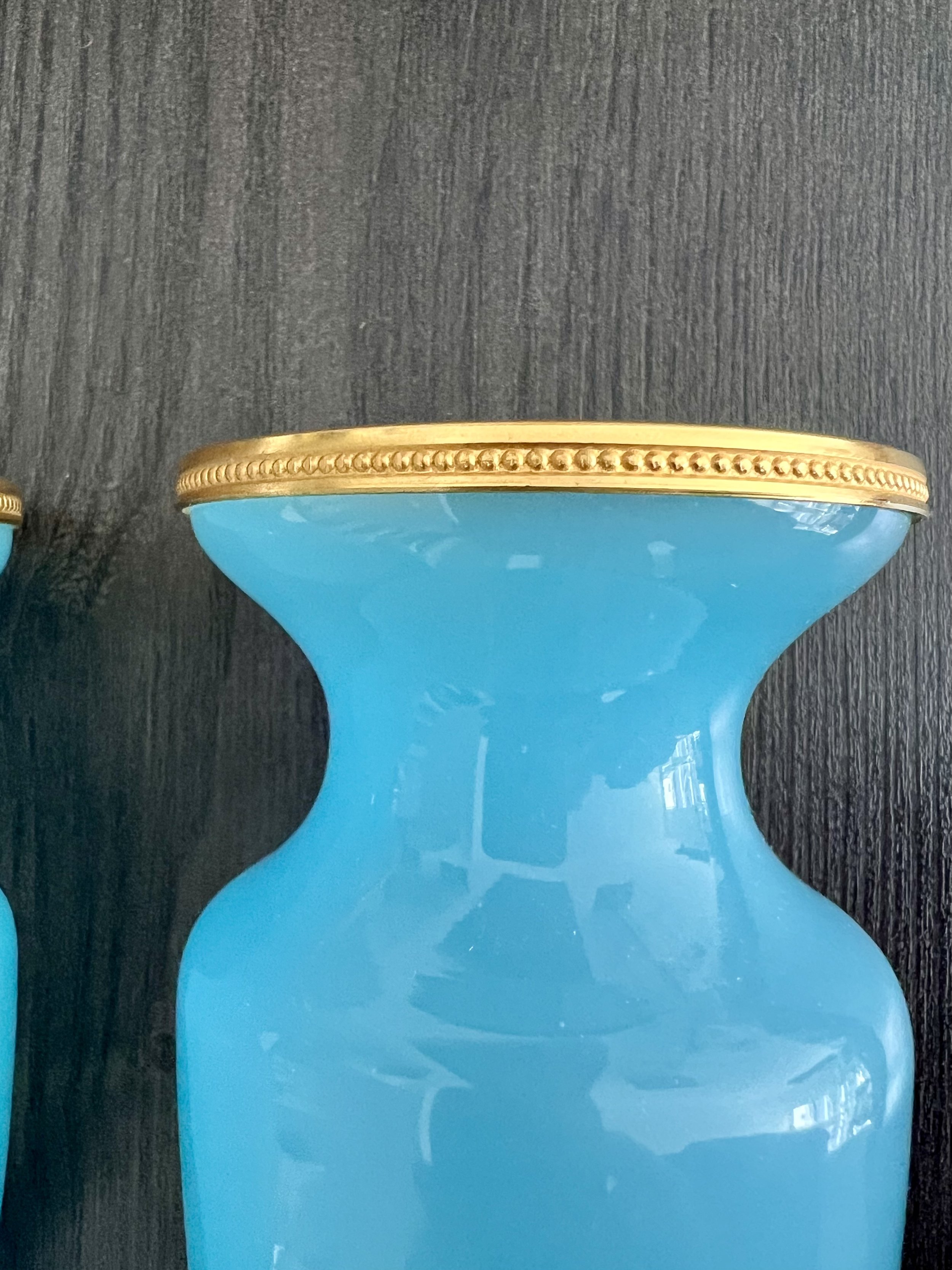 A three-piece antique garniture set made of turquoise blue opaline glass with ornate gilded gold rims, featuring two matching baluster vases and a central cup on a dark wood table.