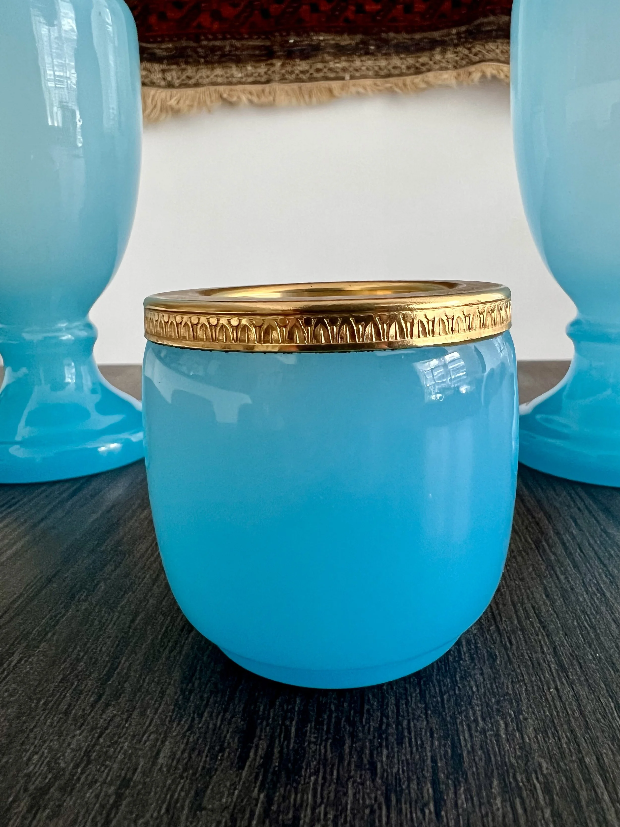 A three-piece antique garniture set made of turquoise blue opaline glass with ornate gilded gold rims, featuring two matching baluster vases and a central cup on a dark wood table.