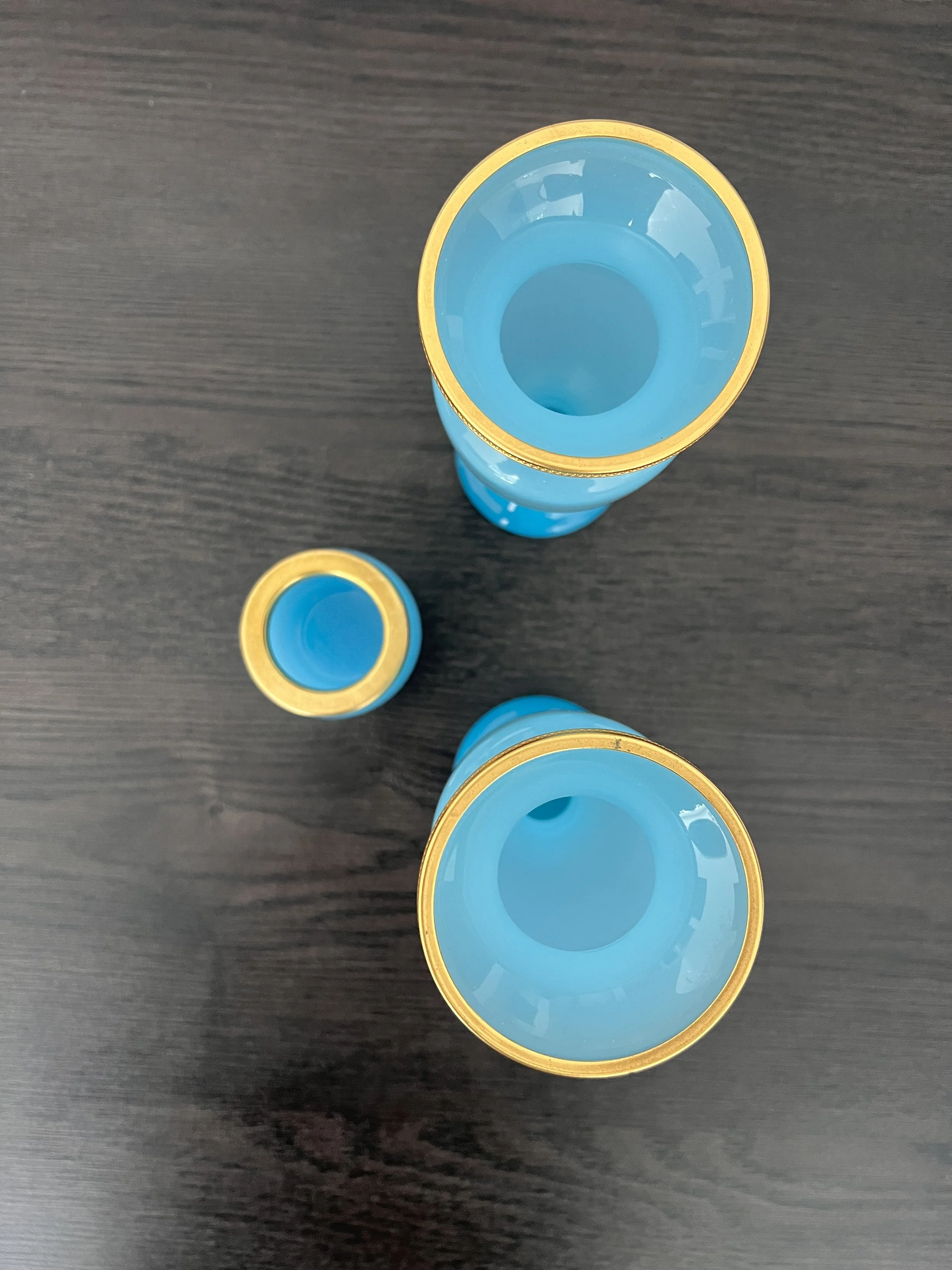 A three-piece antique garniture set made of turquoise blue opaline glass with ornate gilded gold rims, featuring two matching baluster vases and a central cup on a dark wood table.