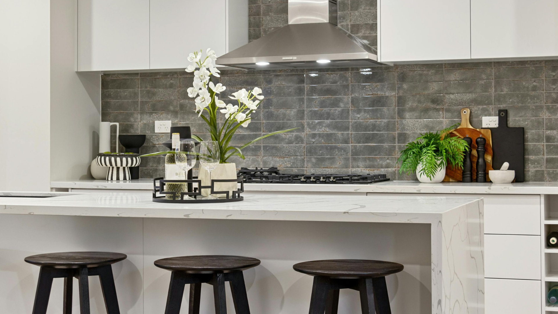 Modern kitchen with gray tiled backsplash, white cabinets, stainless steel range hood, and a white marble island with three black stools. Decor includes a tray with wine glasses and white orchid, a potted fern, and wooden cutting boards.