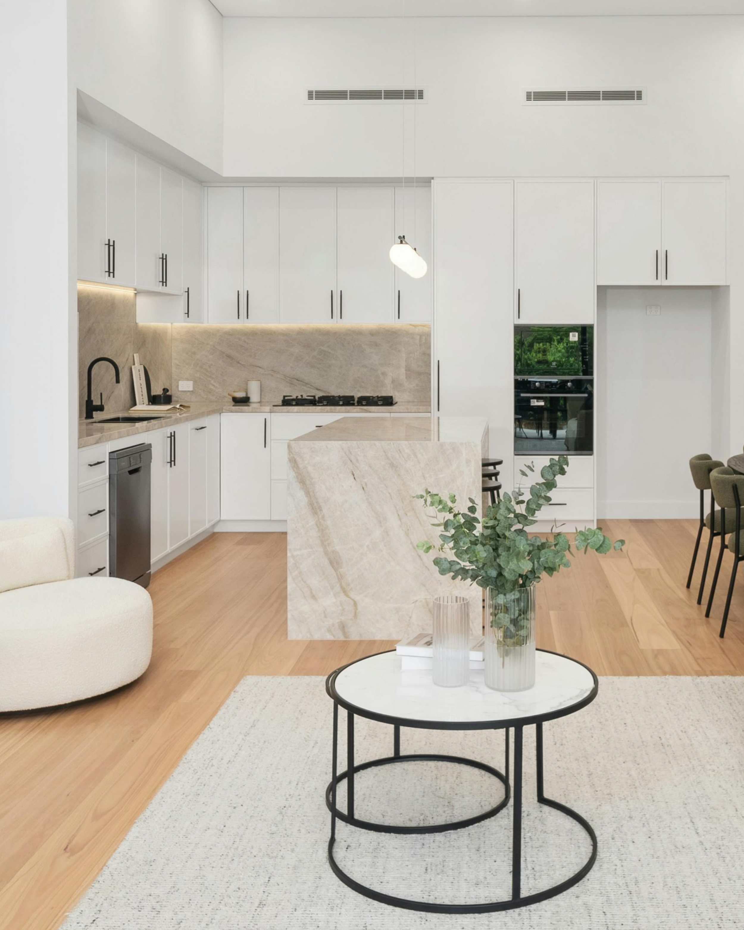 Modern open-plan kitchen and dining area featuring white cabinets, beige countertops and backsplash, built-in oven, integrated rangehood and microwave, black faucet, round dining table.