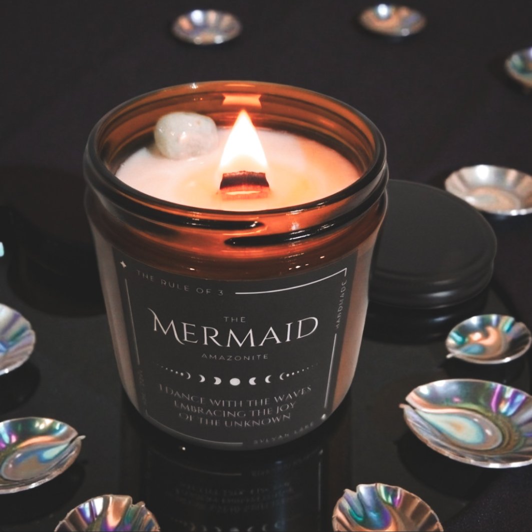 “Mermaid Amazonite ritual candle burning with a crackling wood wick, surrounded by seashells on a dark reflective surface.”