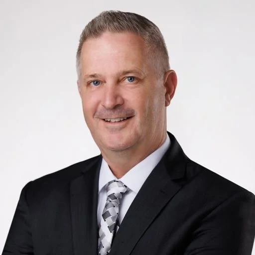 A professional headshot of a man with short, light-colored hair, wearing a black suit, white shirt, and patterned tie, smiling against a plain white background.