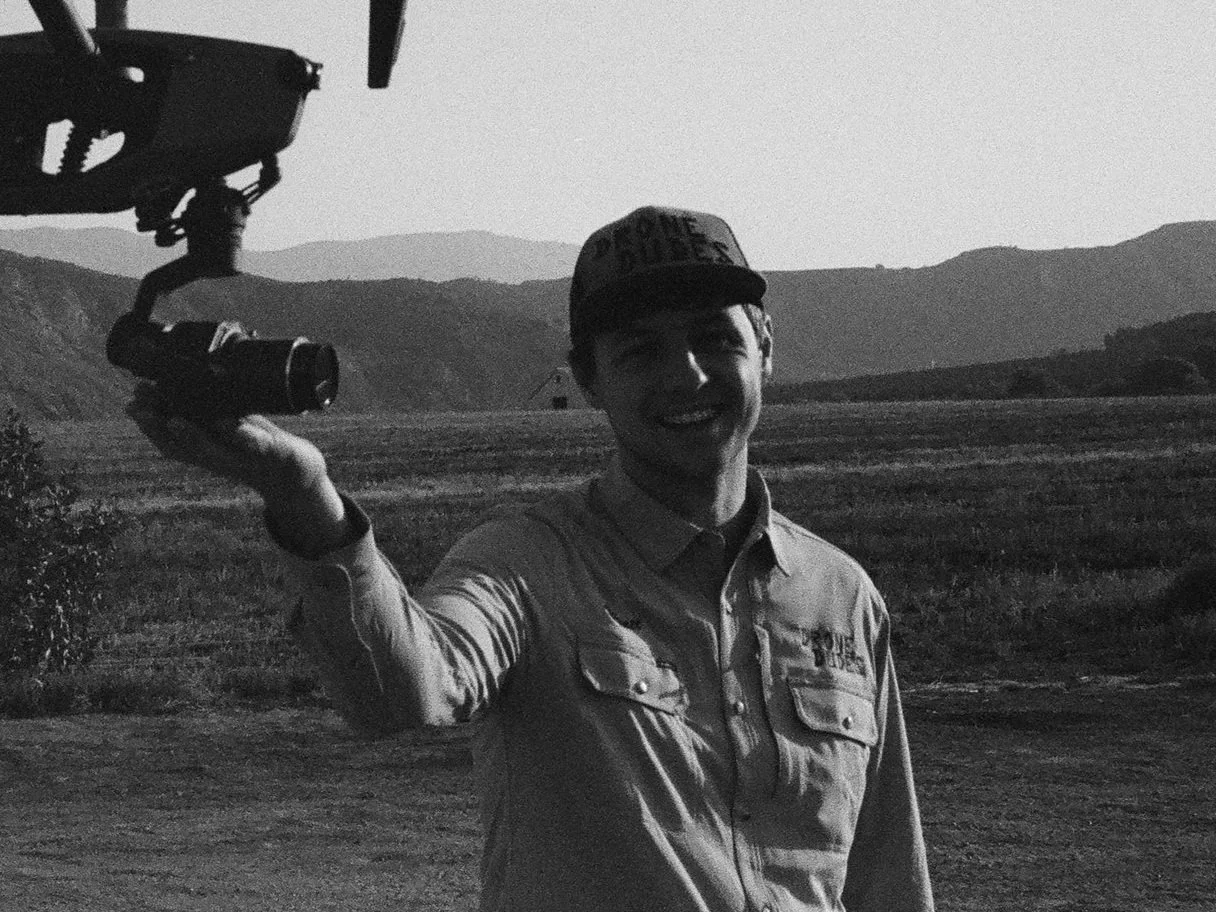 A man smiling and holding a drone controller outdoors in a rural area with mountains in the background.