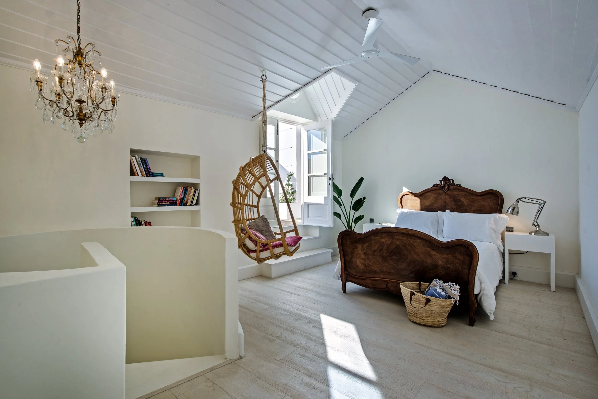 Bright bedroom with white walls and ceiling, wooden bed with white bedding, white bedside table with reading lamp, hanging egg chair with red cushion, open window with sunlight, potted plant near window, chandelier and ceiling fan.