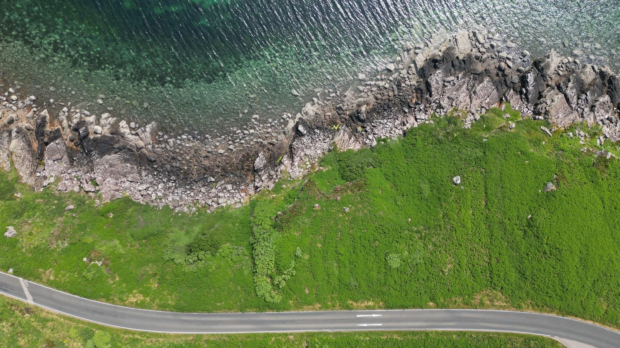 An aerial view of a green grassy landscape with large rocks and a road running along the bottom, adjacent to a body of water with a rocky shoreline.