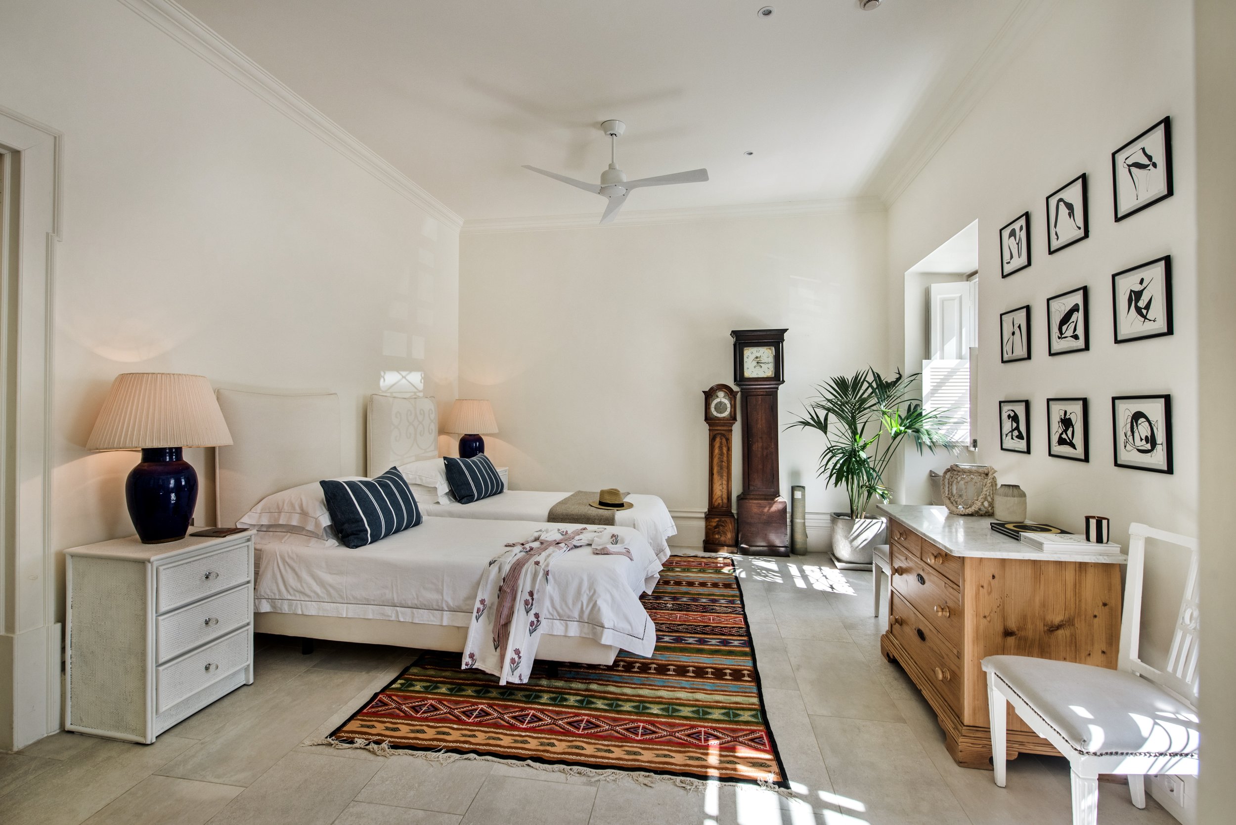 A spacious hotel room with two beds, white bedding, striped pillows, a nightstand with a lamp, a colorful patterned rug, a chandelier, a tall clock, a potted plant, a wooden dresser, a white chair, and framed black and white artwork on the wall.