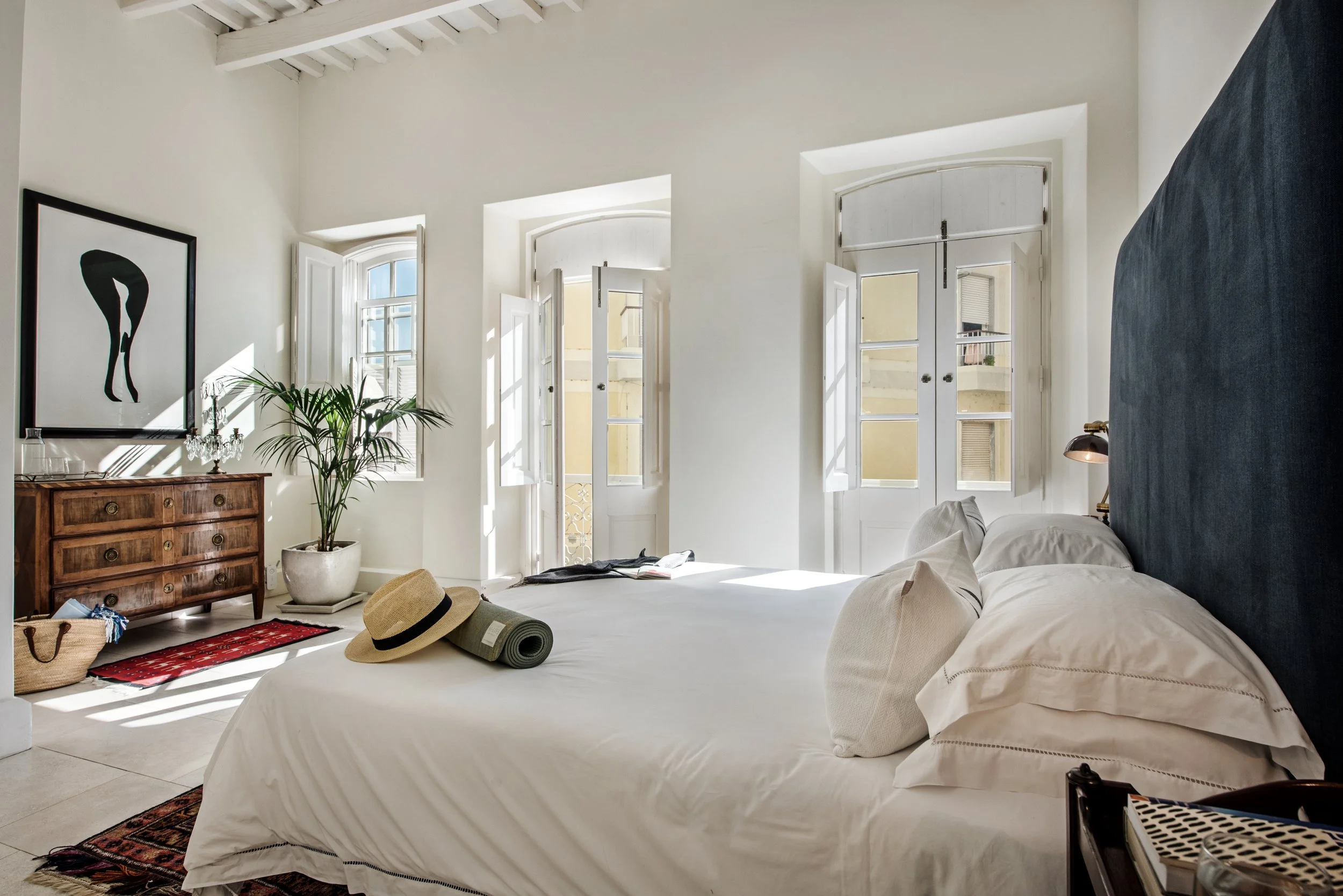 Bright bedroom with white walls, multiple open windows letting in sunlight, a large bed with white linens and pillows, a black headboard, a straw hat, a rolled-up mat, a potted plant, a wooden dresser with art above, and various bags on the floor.