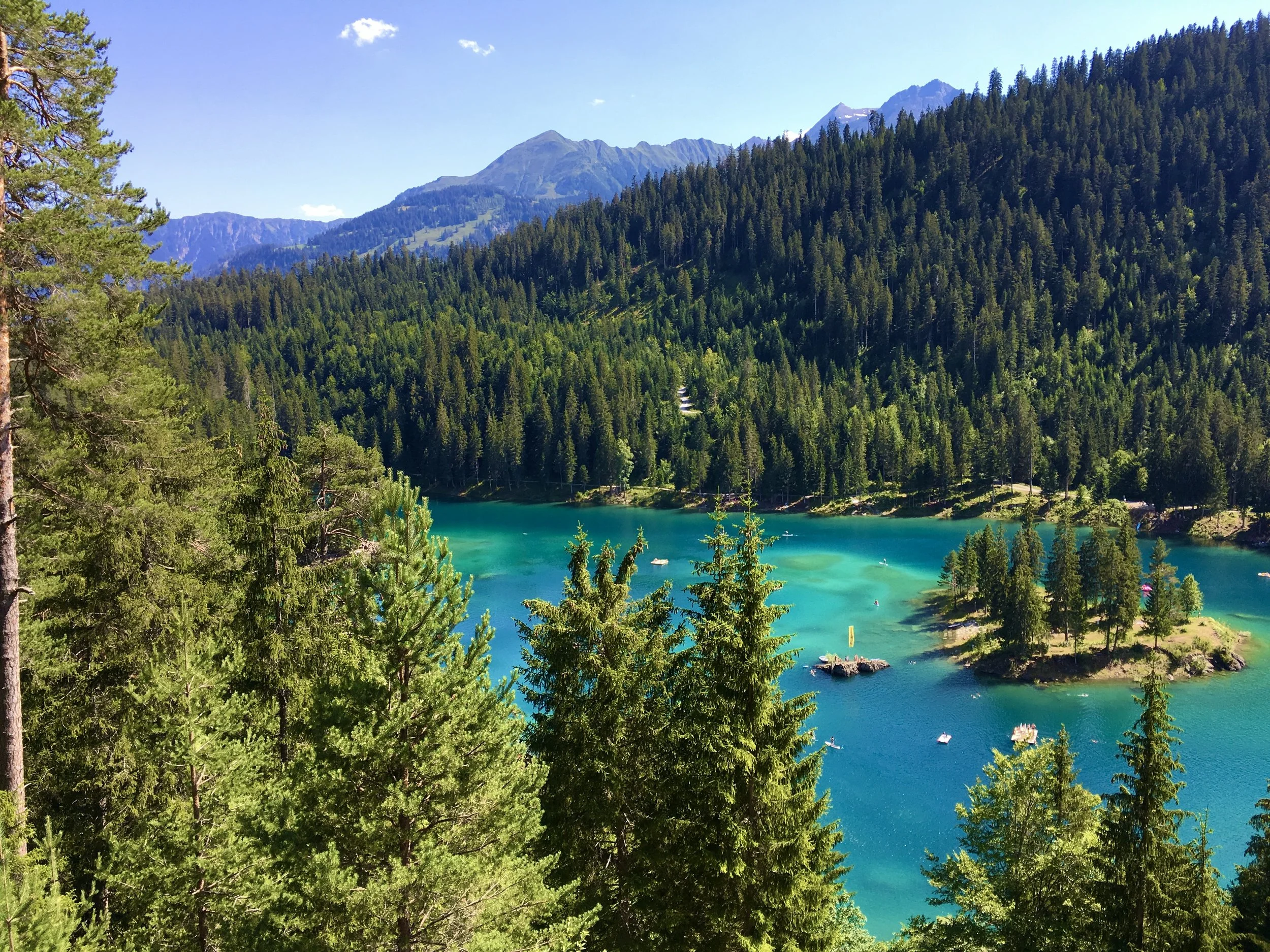 A scenic view of a turquoise lake surrounded by dense evergreen trees with a mountain range in the background under a clear blue sky.
