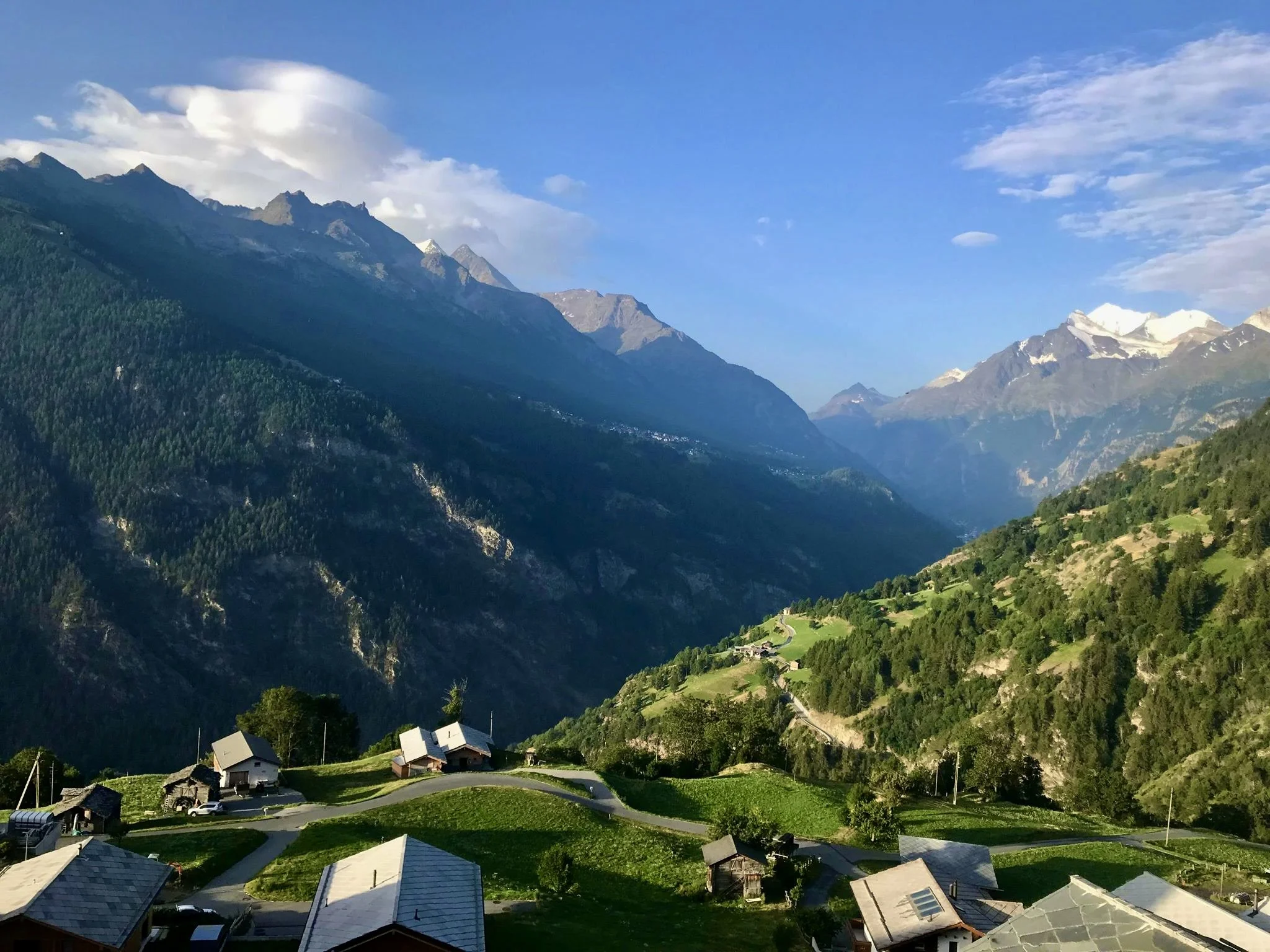 Mountain landscape with a village in the foreground, lush green terraces, winding roads, and snow-capped peaks under a partly cloudy sky.