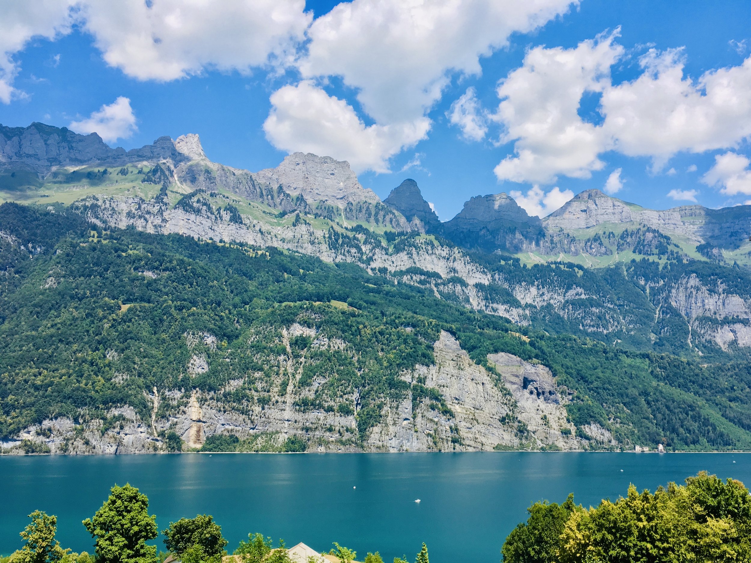 Scenic view of a mountain range with rocky peaks and green slopes, beside a calm lake reflecting the sky, with trees in the foreground and a partly cloudy blue sky above.