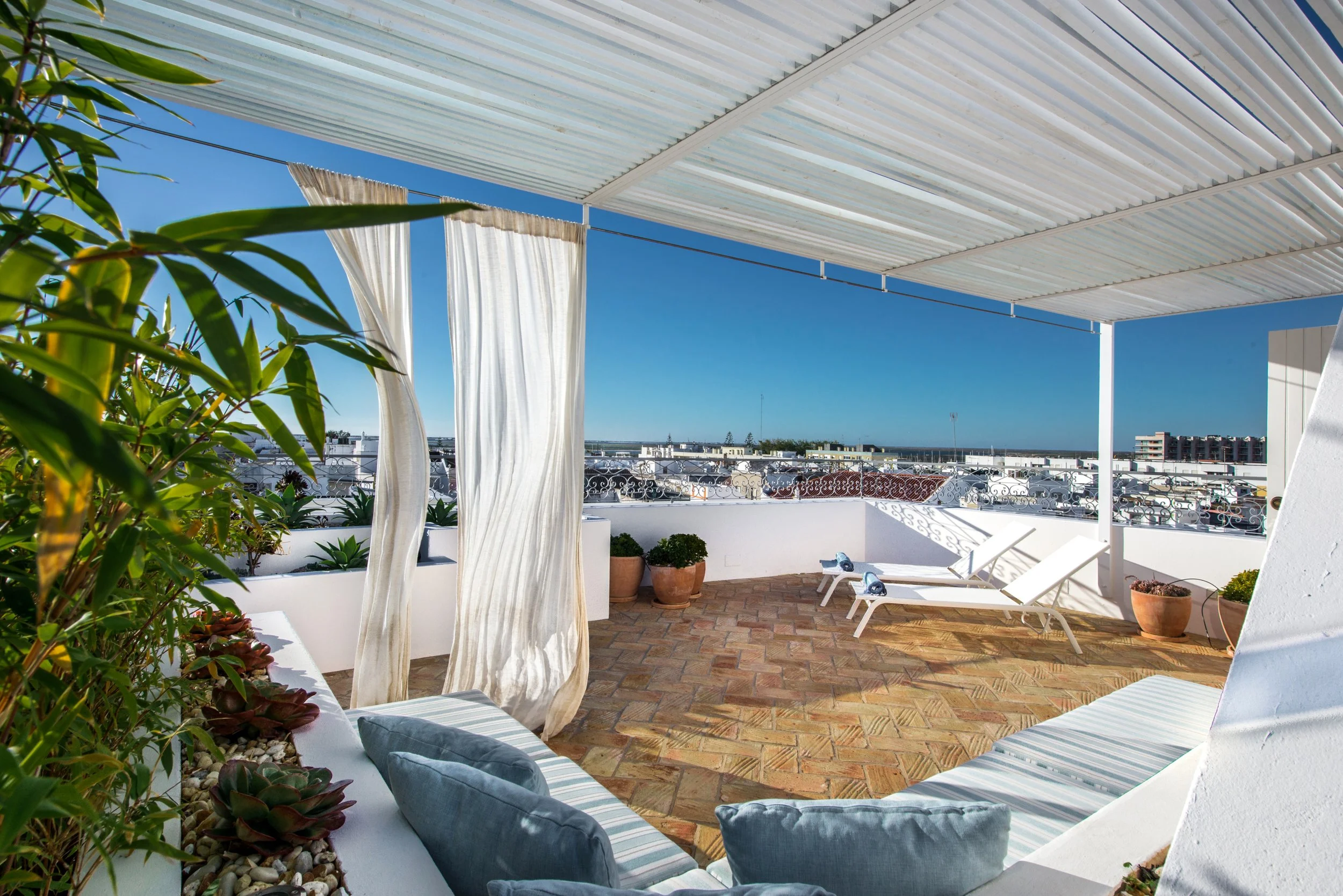 A rooftop terrace with white curtains, potted plants, and lounge chairs under a white roof, overlooking a cityscape on a sunny day.