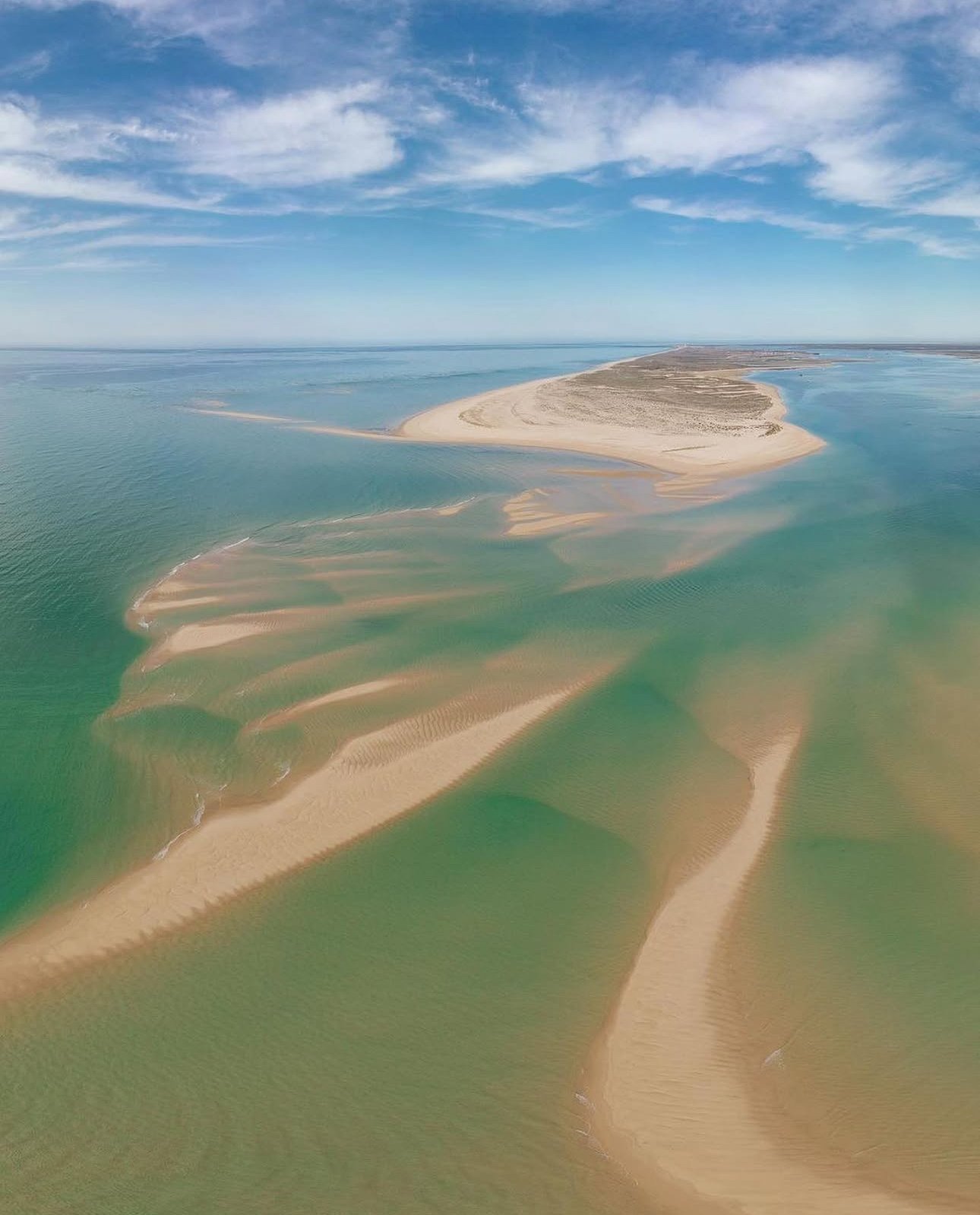 Aerial view of sandbars and shallow water formations in a coastal area with blue sky and wispy clouds.