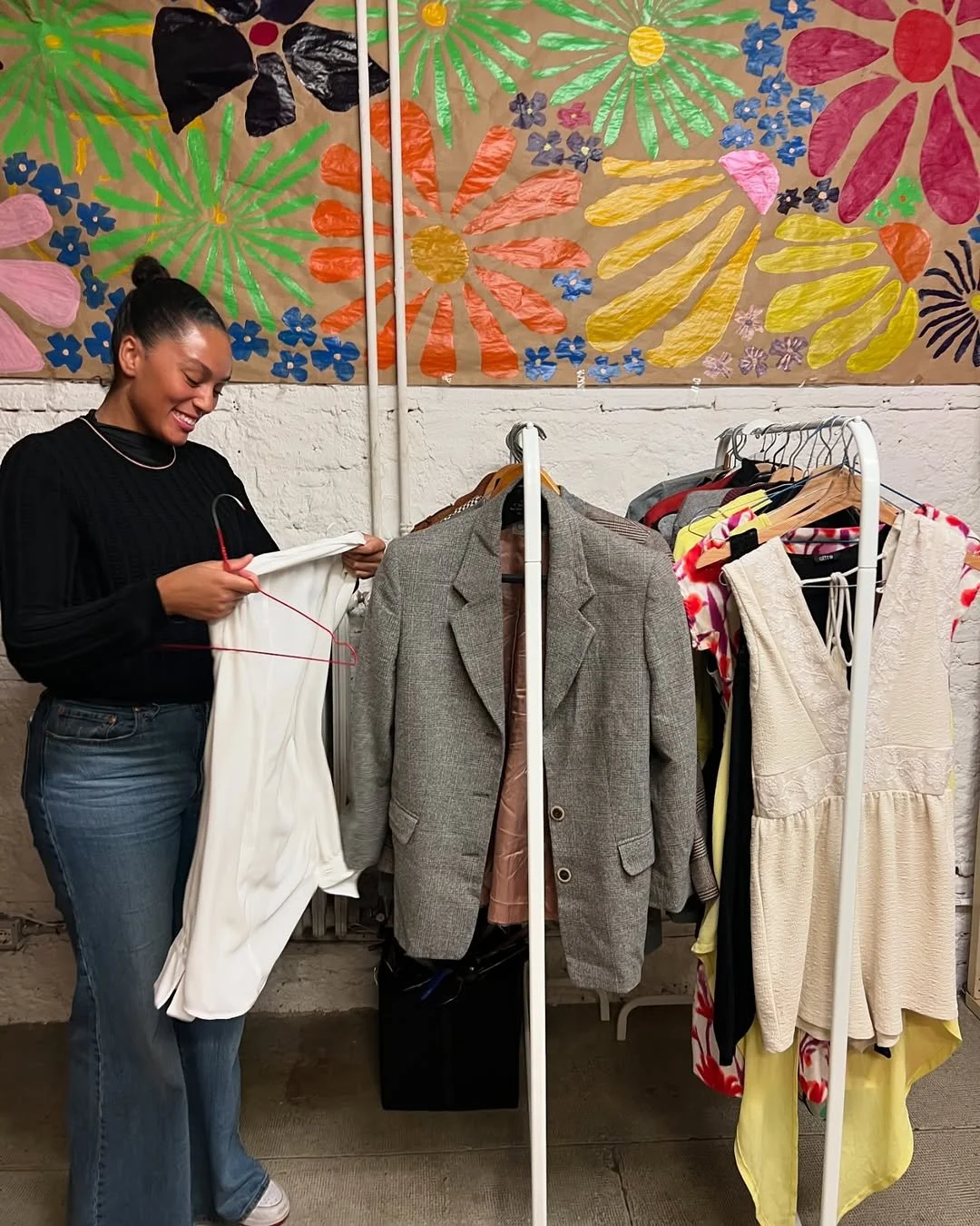 A woman with a bun hairstyle wearing a black sweater and jeans shopping for clothes at a store, smiling as she holds a white piece of clothing on a hanger. She is standing next to a clothing rack with a gray blazer, and a colorful abstract floral artwork is displayed on the wall behind her.