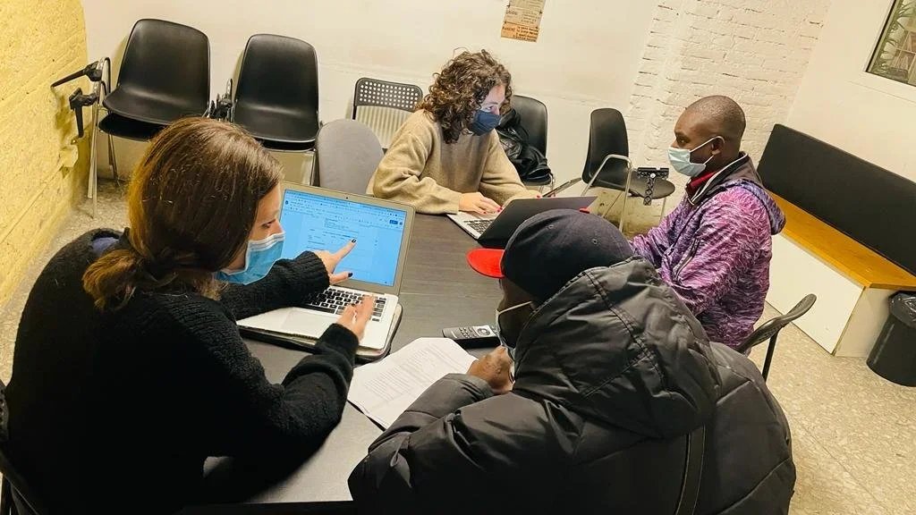 Four people wearing face masks sitting around a table, using laptops and discussing in a small meeting room.