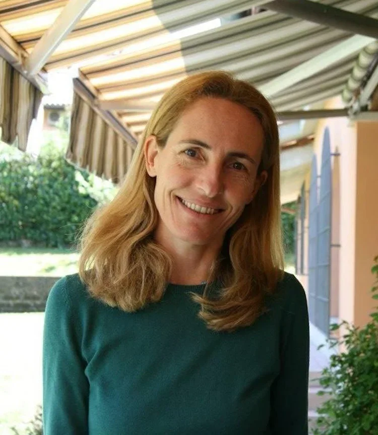 A woman with long blonde hair wearing a teal top standing outdoors under a striped awning, smiling at the camera.