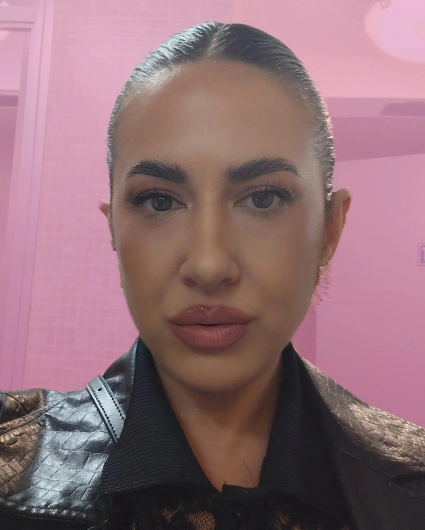 Woman with dark hair slicked back in front of a pink background