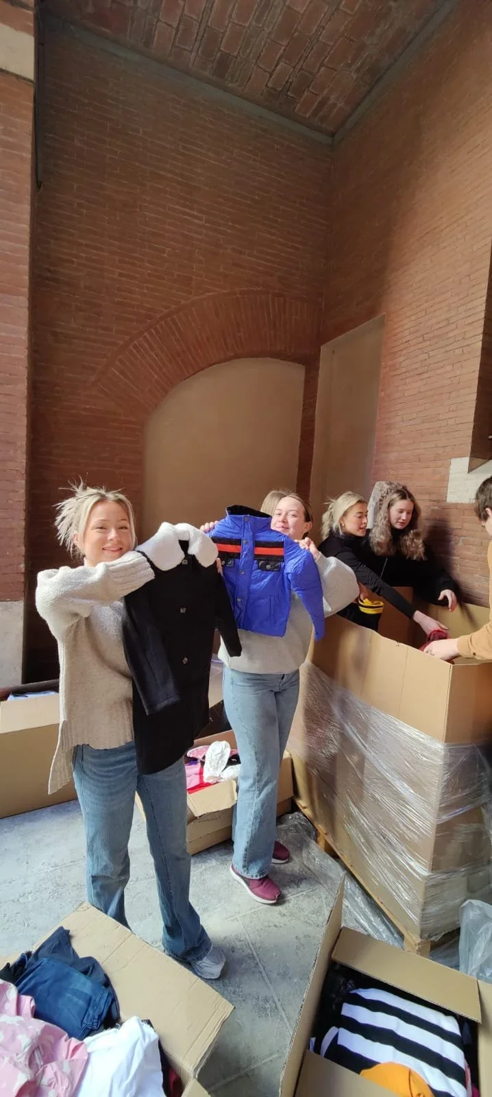 Two smiling women holding up shirts while others are packing or sorting clothes in a room with brick walls.