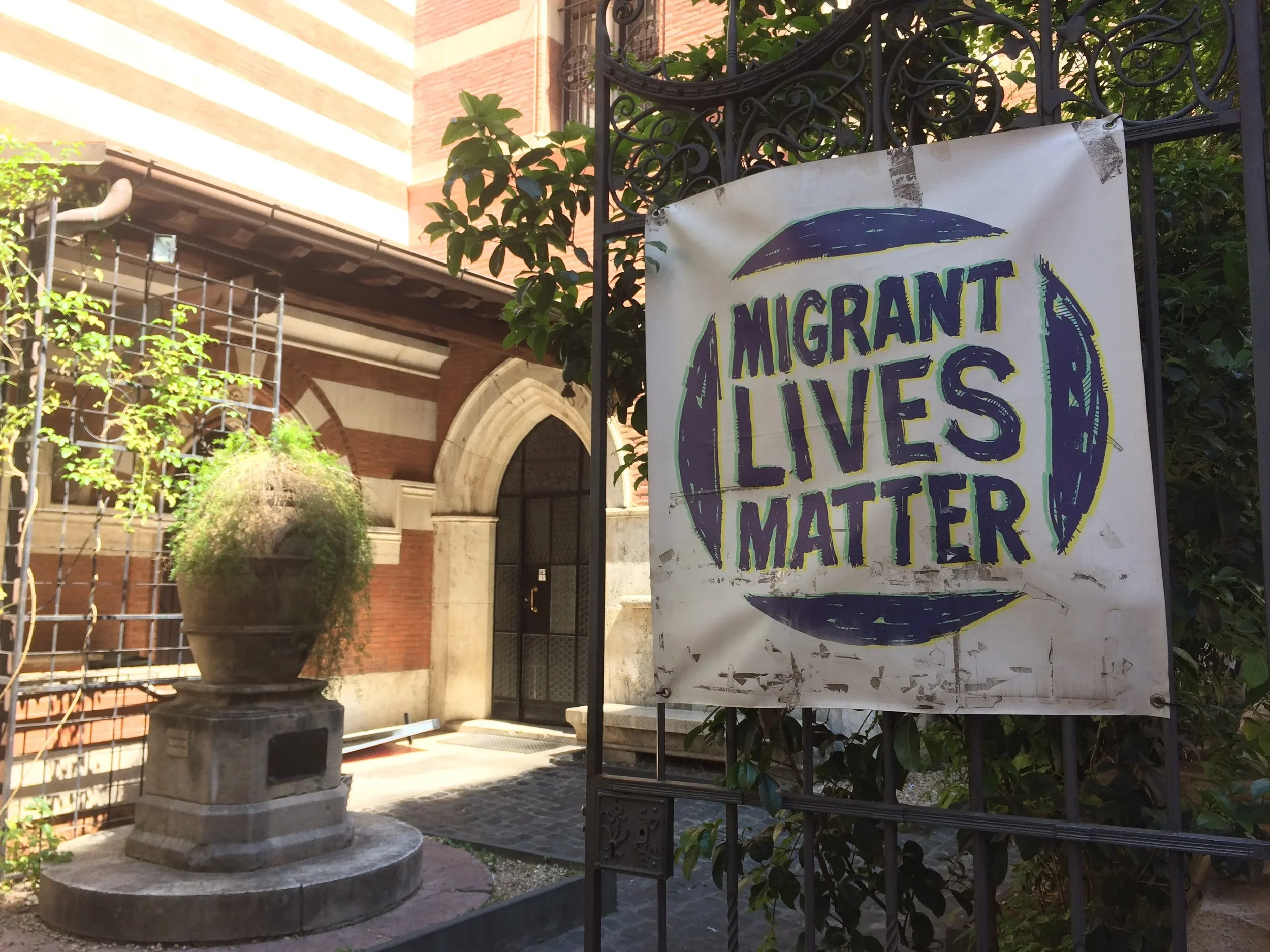 A sign with the text 'Migrant Lives Matter' attached to a black wrought iron fence outside a building with a stone archway entrance and a brick facade, with a large plant in a decorative stone planter nearby.