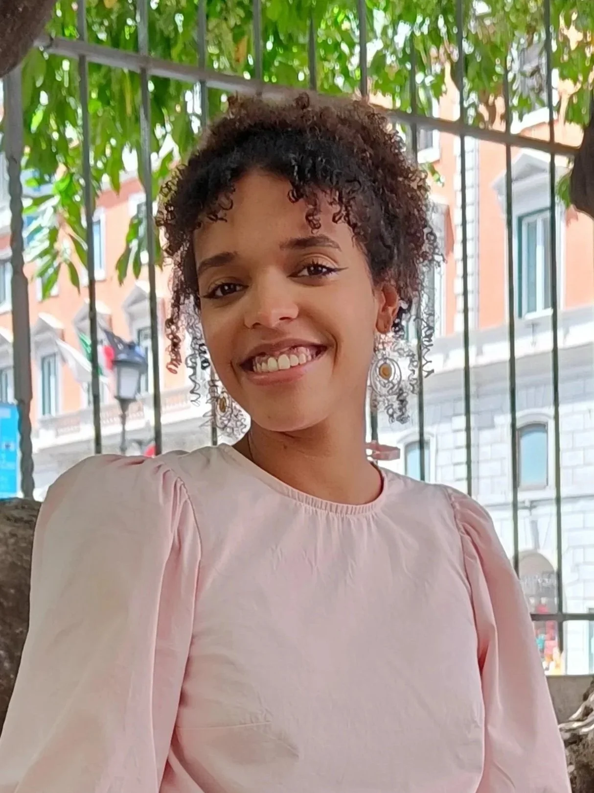 Woman in a light pink shirt with curly hair in an updo, with a tree and metal fence in the background
