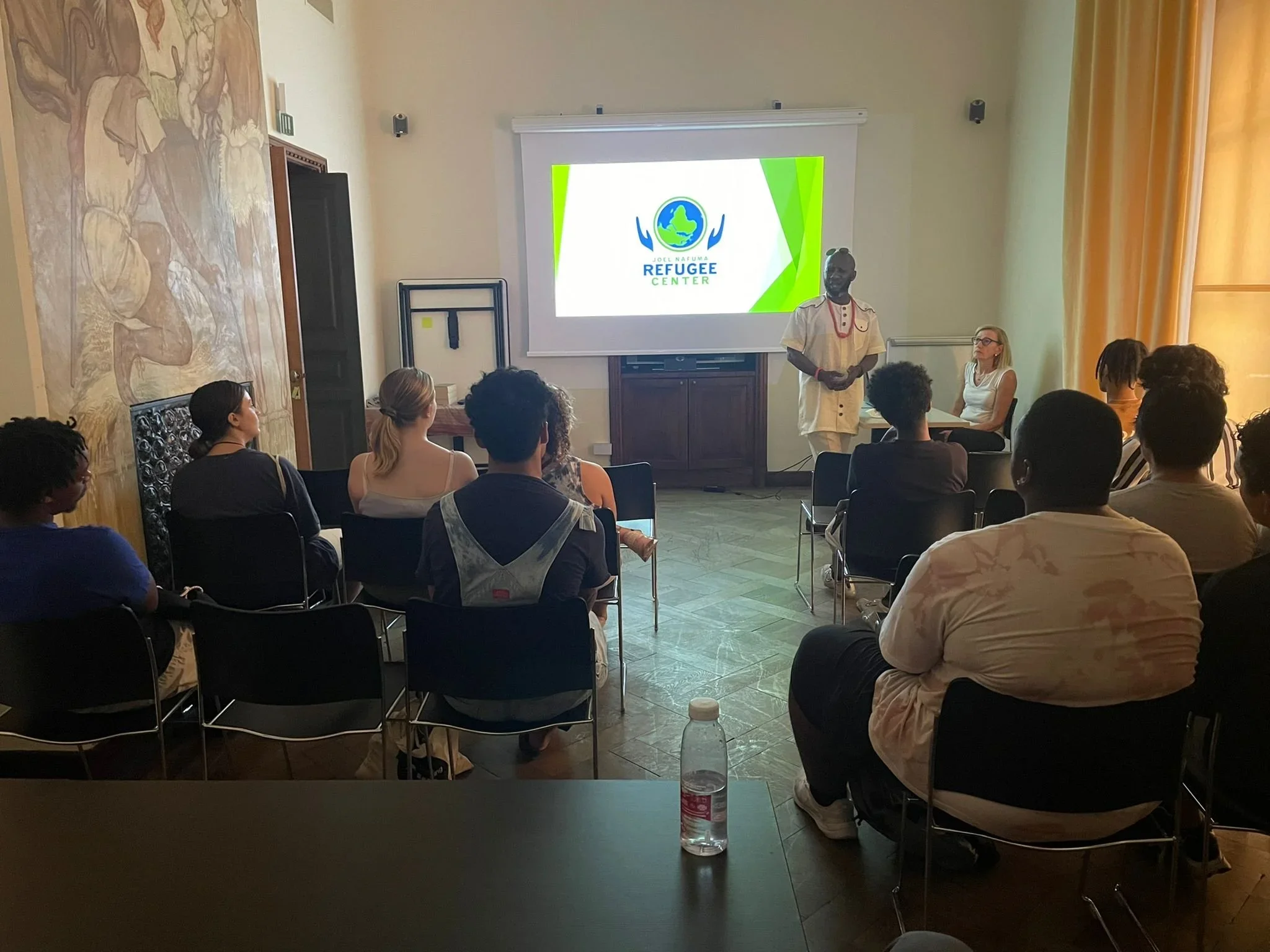 A group of diverse people seated and listening to a speaker in a room, with a projector screen displaying a logo for the Refugee Center.