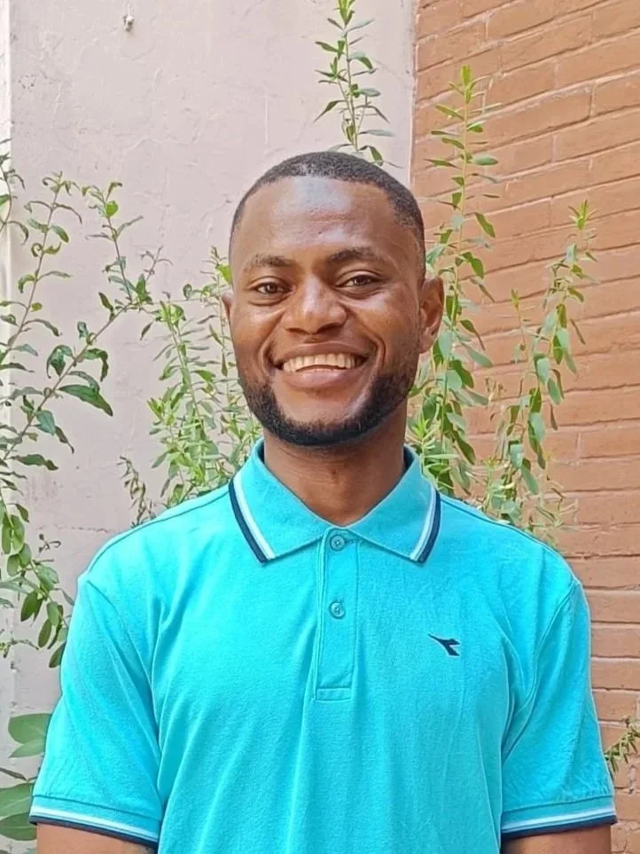 A smiling man with short hair and a beard wearing a blue  t-shirt, standing against a brick wall.