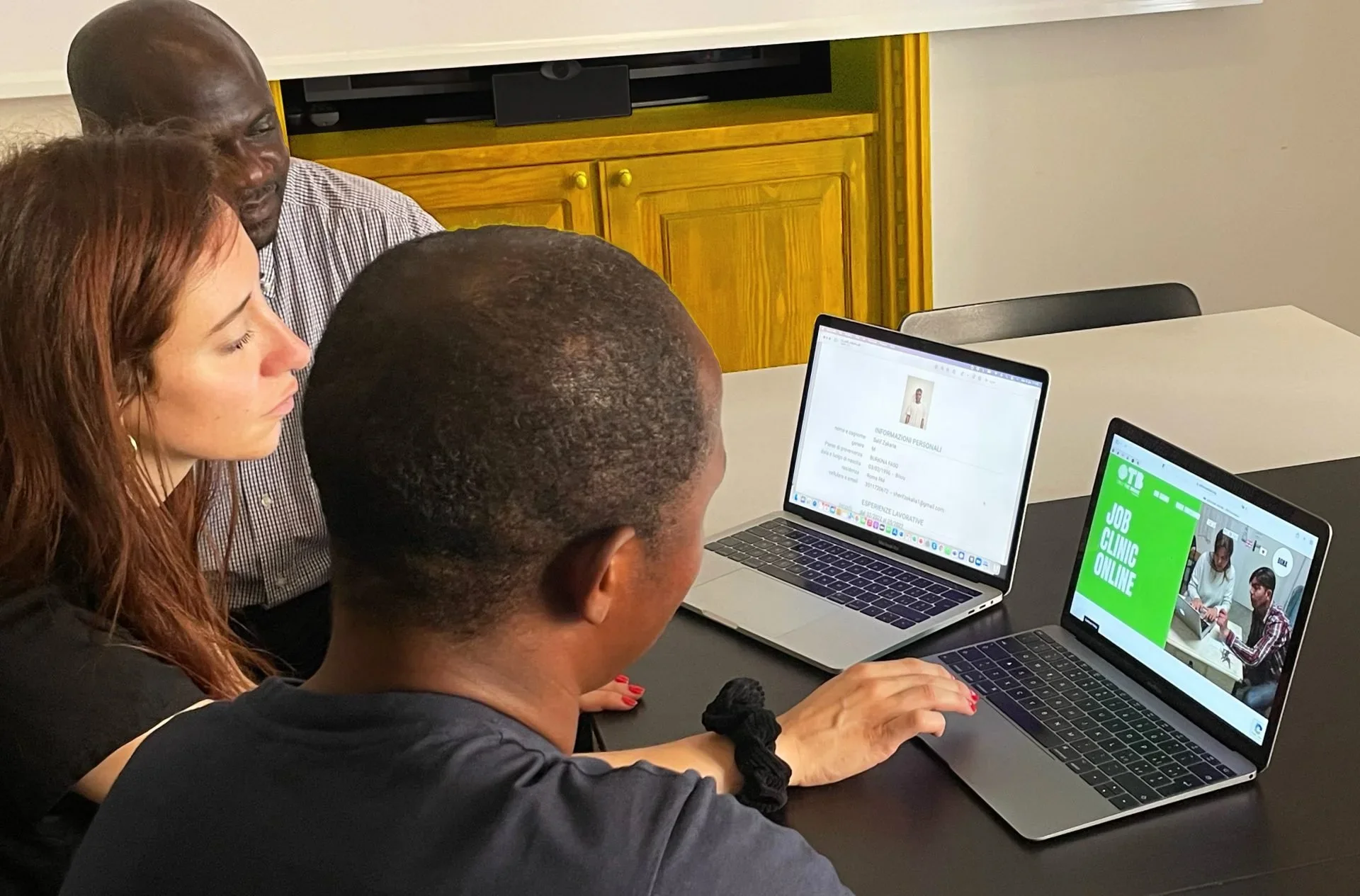 Three people sitting at a table working on laptops with a wooden cabinet in the background.