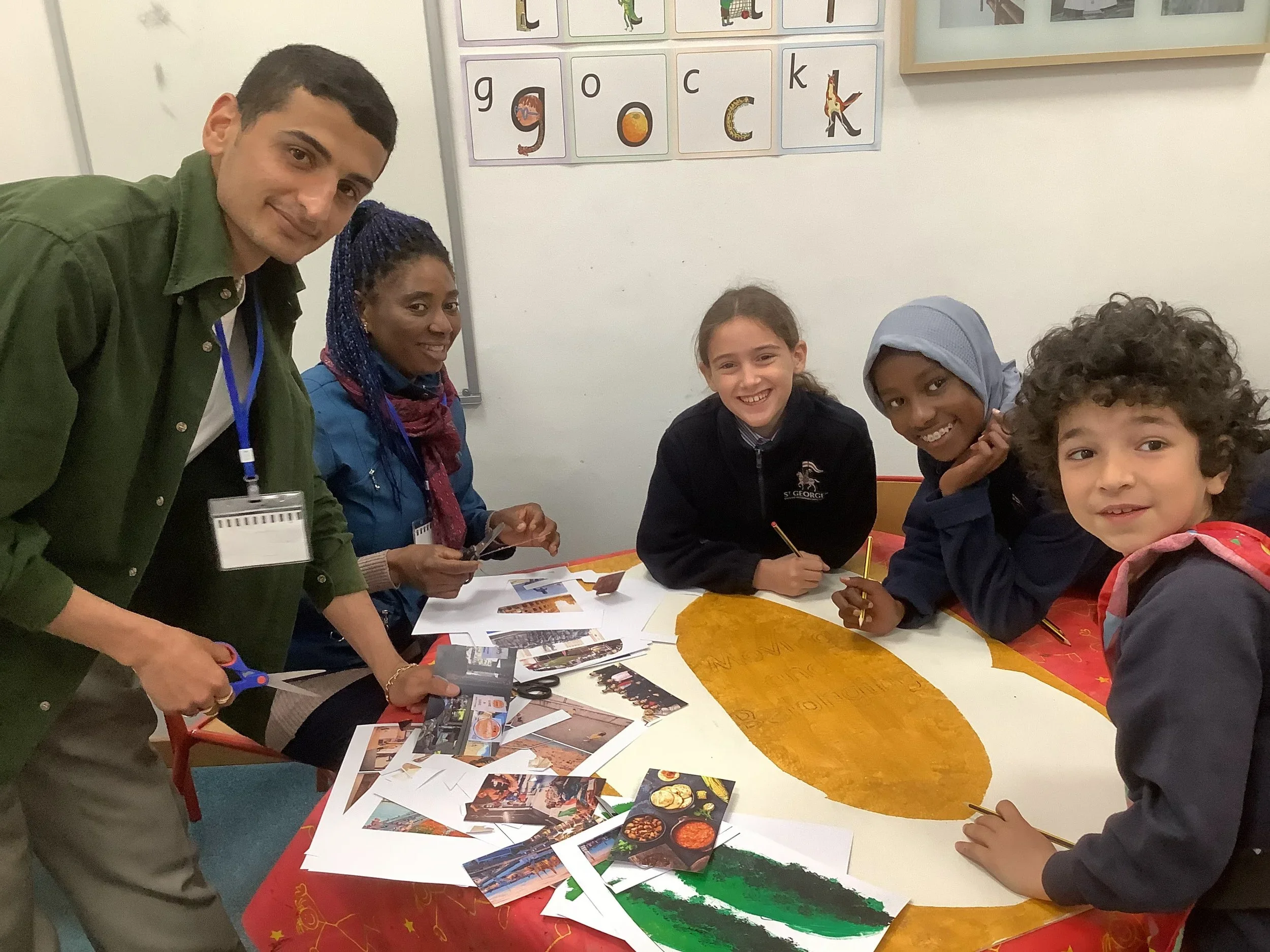 Group of five people, two adults and three children, gathered around a table in a classroom, engaging in arts and crafts activities. The table has photos, colored papers, and art supplies. Kids are smiling and working on a large, colorful poster.
