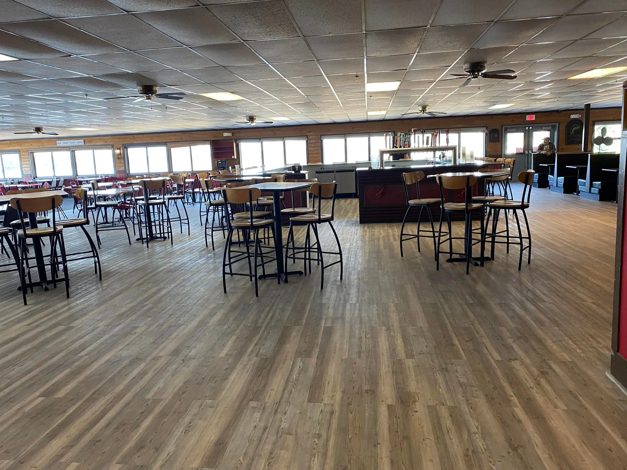 Empty restaurant dining area with tables and chairs, large windows, and a bar in the background.