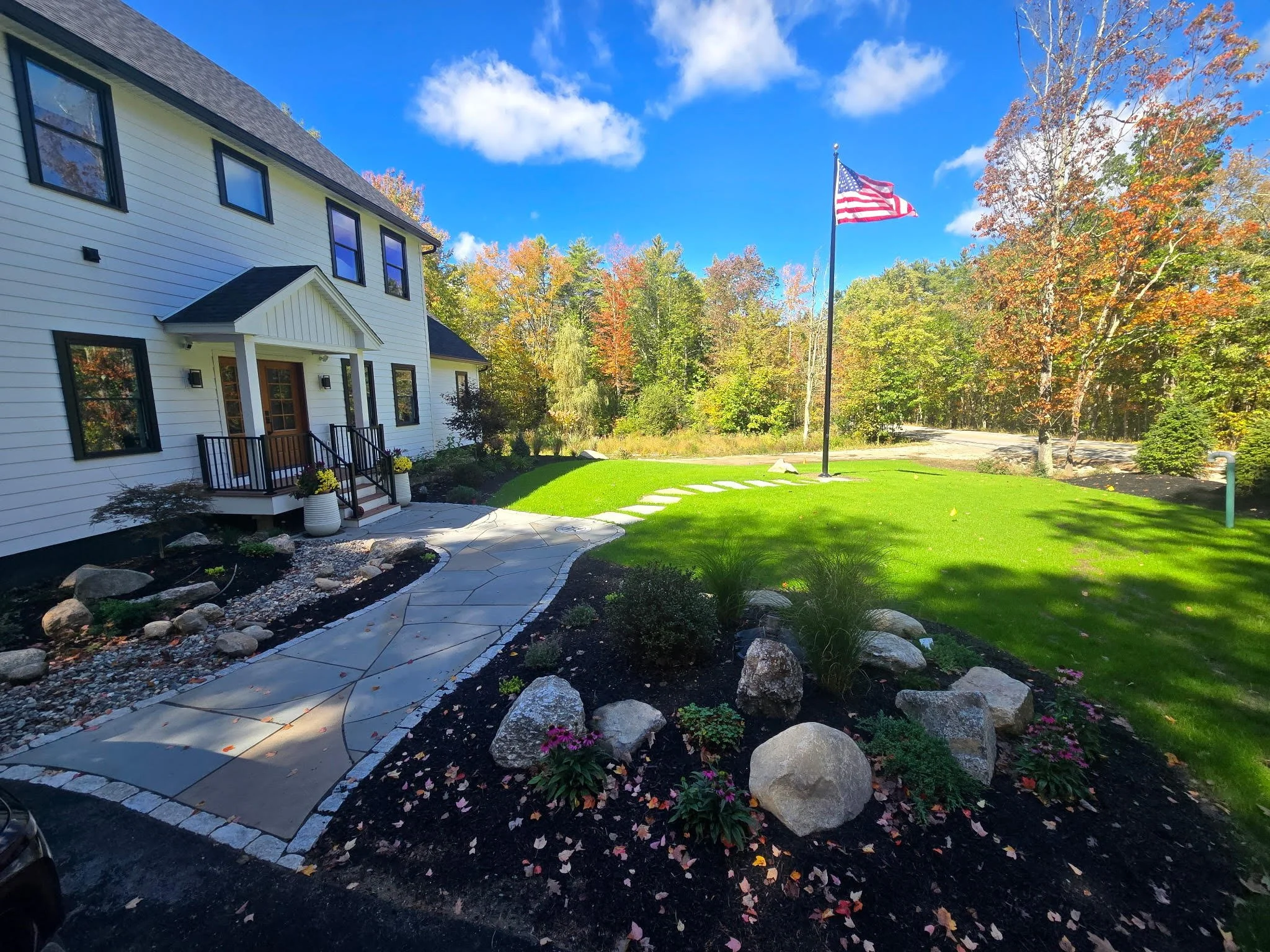 A white house with black window frames and a small front porch with plants in pots. A stone walkway leads to the porch, and the yard features green grass, trees with fall foliage, and a large American flag on a pole. The sky is blue with some clouds.
