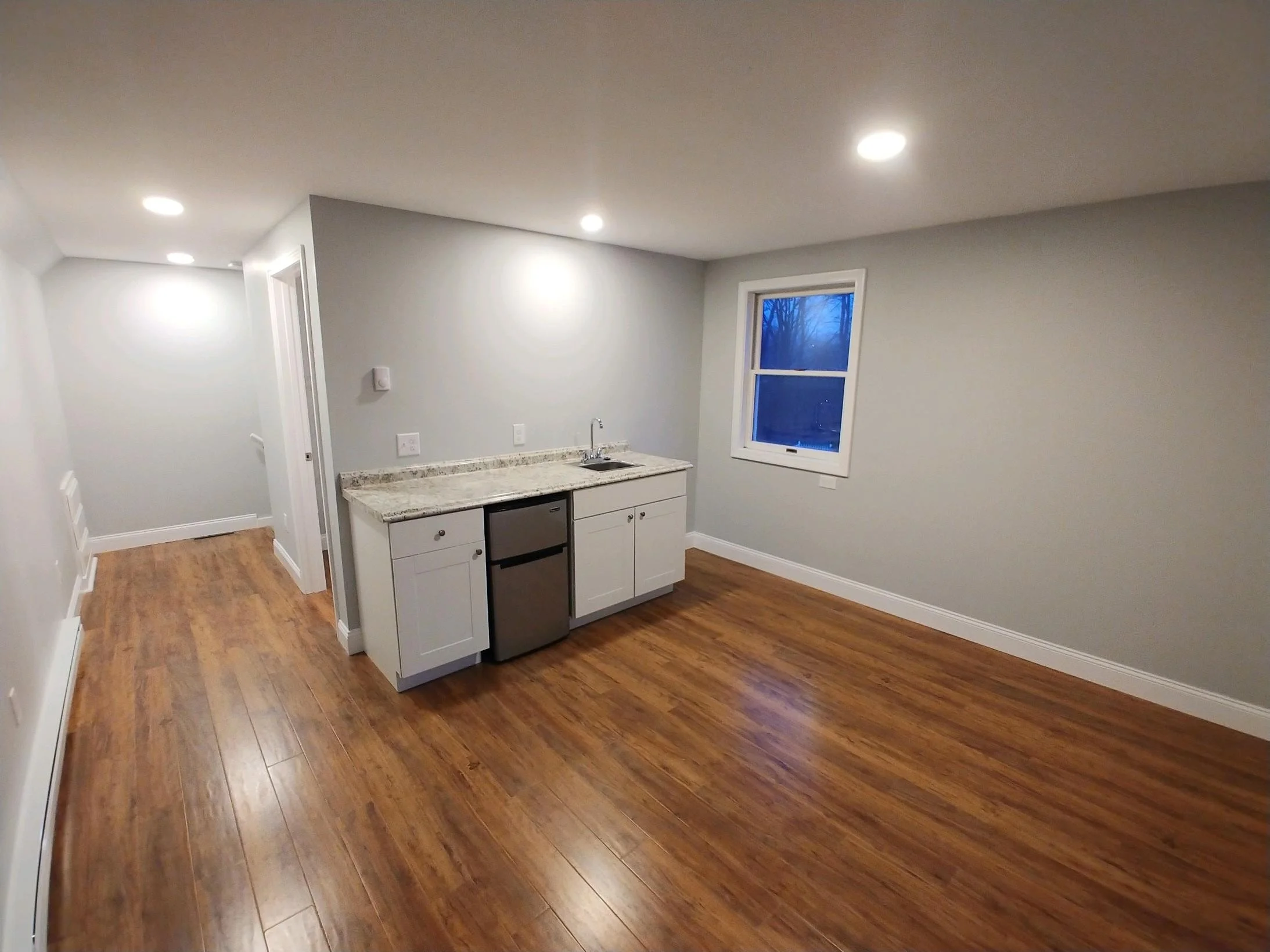 Empty living space with hardwood floors, a small kitchen with white cabinets, granite countertop, and a window showing a blue evening sky.
