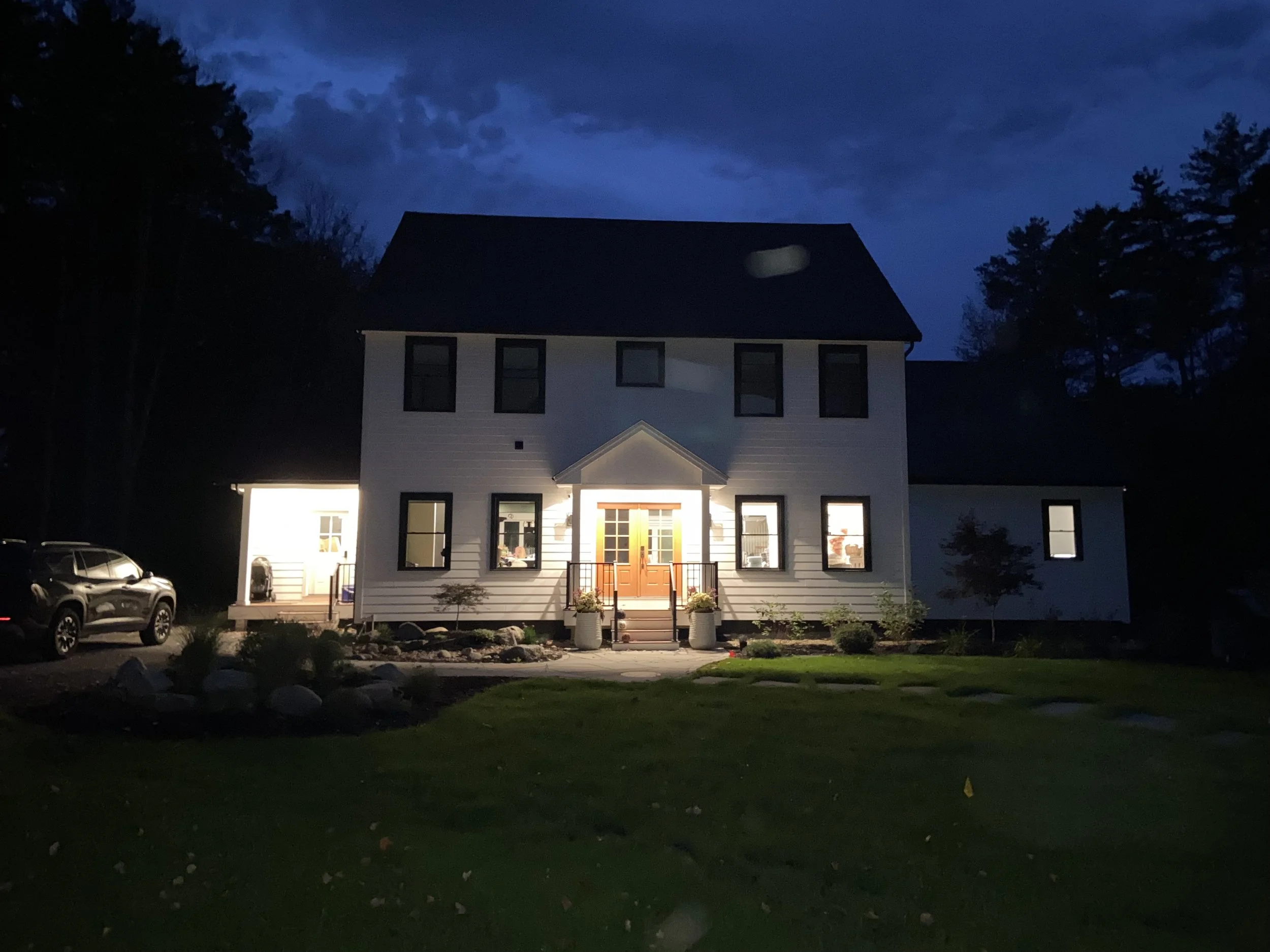 A two-story white house with black window frames and a front porch, illuminated at night, with a dark sky in the background and a driveway with a parked car.