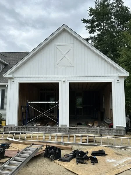 Under construction garage with two open bays, tools, a ladder, and building materials on the ground.