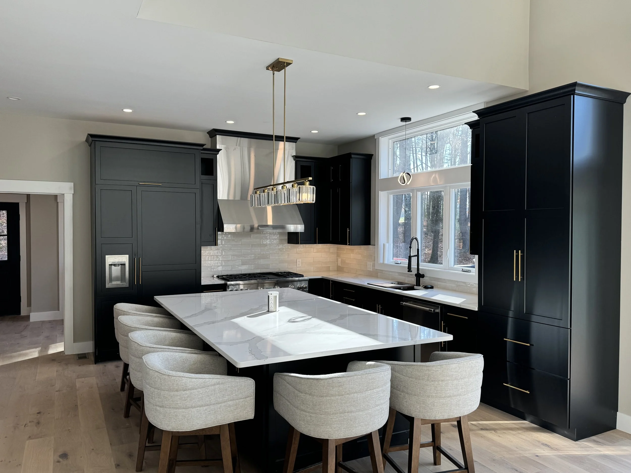 Modern kitchen with black cabinets, a white marble island, light-colored chairs, and large window with view of trees.