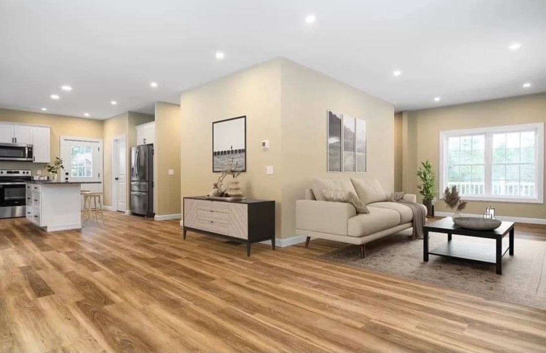 Open-concept living room and kitchen with hardwood floors, beige walls, white cabinetry, stainless steel appliances, a beige sofa, coffee table, and large window.